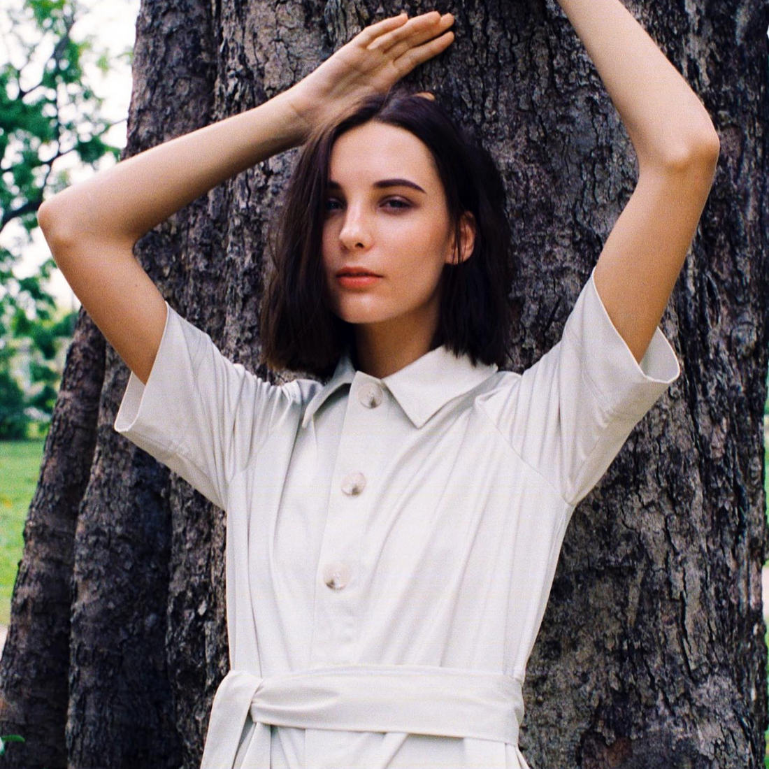 A woman with short brown hair leans against a tree. She has her arms raised above her head. She is wearing a white shirt dress.