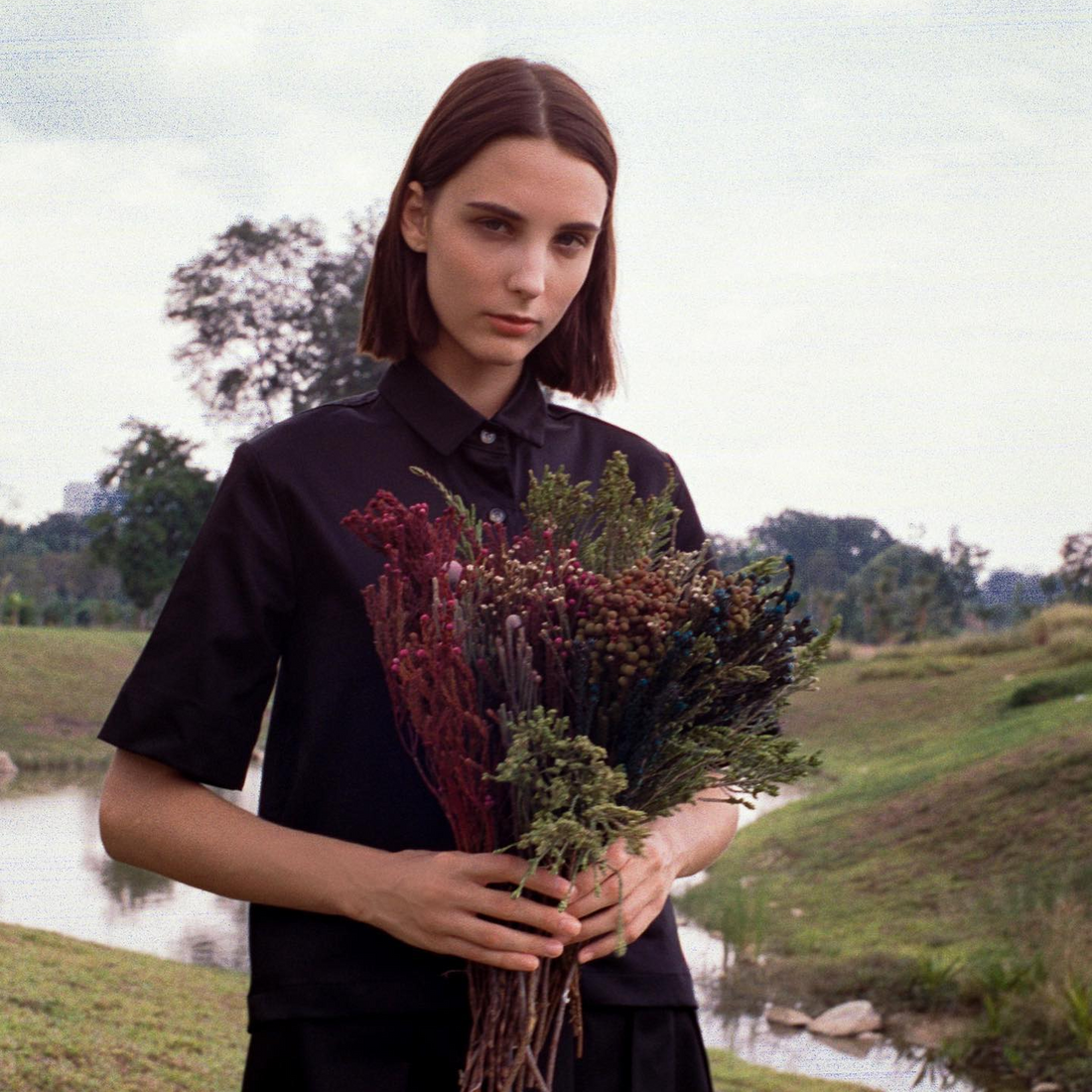 A woman with short dark brown hair stands in a field. She is wearing a black shirt dress and is holding a bunch of flowers