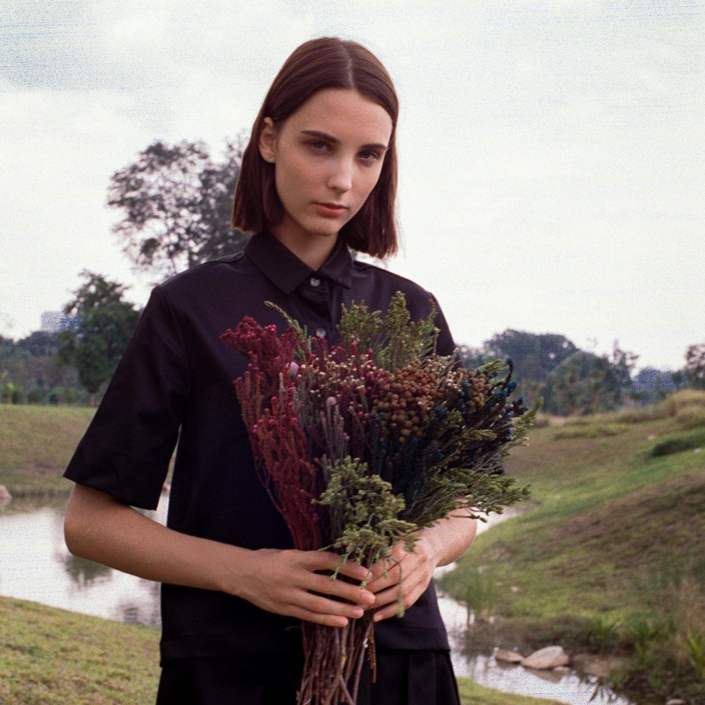 A woman with short dark brown hair stands in a field. She is wearing a black shirt dress and is holding a bunch of flowers