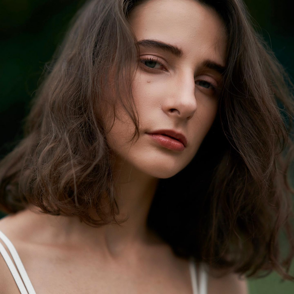 Close up of a woman face. She has dark brown shoulder length hair and is wearing a top with white straps