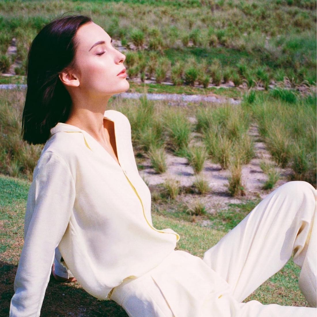 A woman with dark brown hair sits on some grass. She is wearing a white shirt and trousers and has her eyes closed.