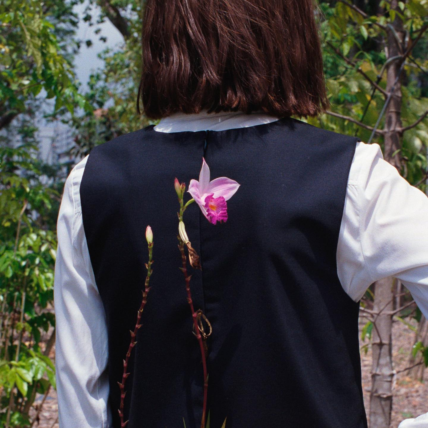 A woman with short brown hair has her back to the camera. She is standing in a forest. She is wearing a white shirt and navy blue waistcoat. She is holding a pink flower behind her back.