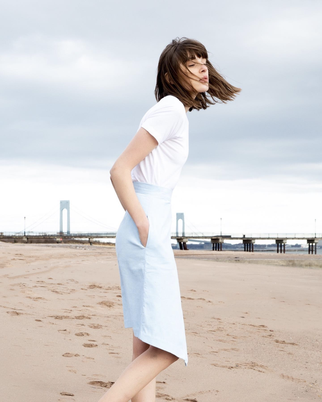 A woman with short dark brown hair wearing a blue skirt and white t-shirt stood on a beach