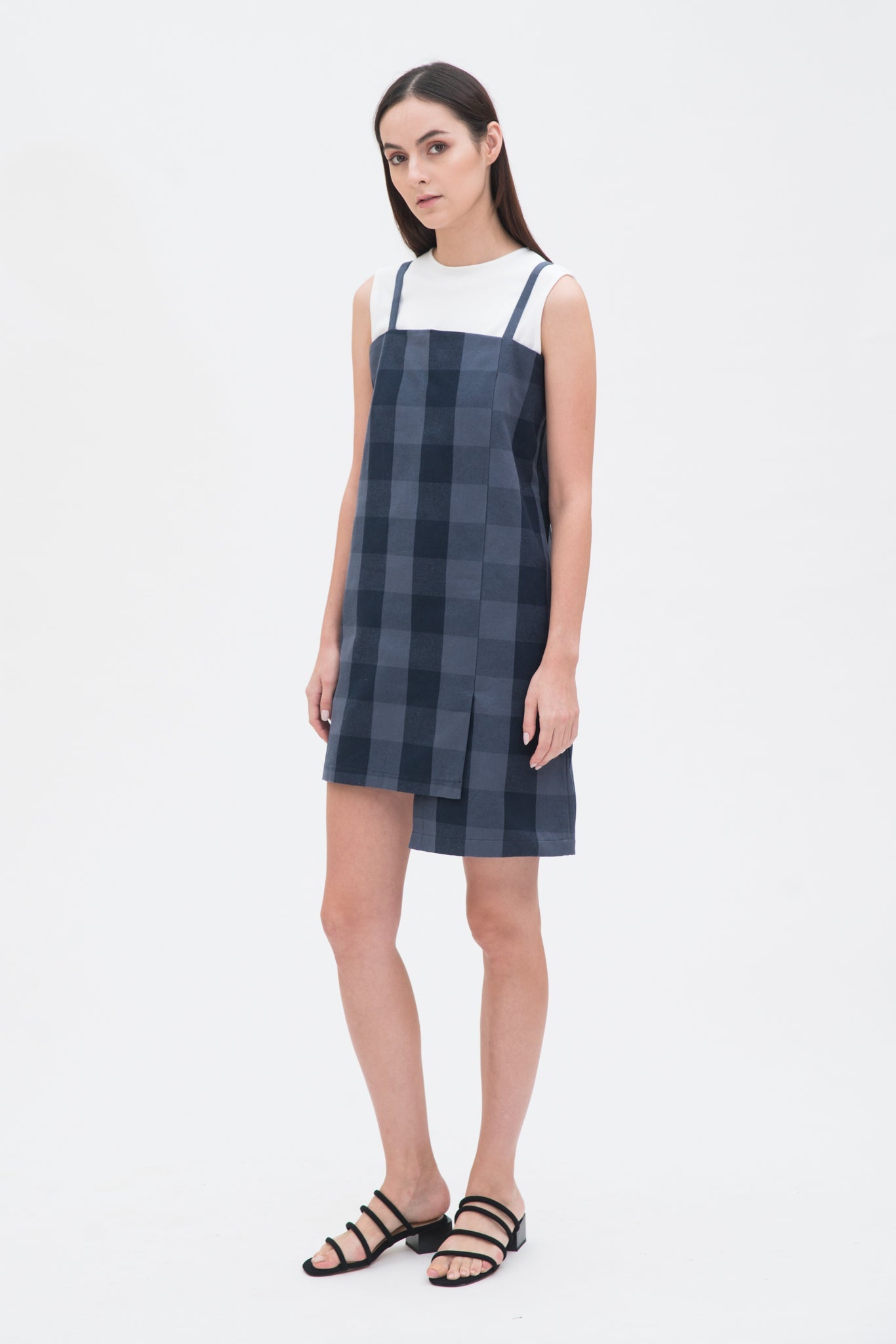 A woman with long dark hair is wearing a navy check dress with a white top underneath and  black sandals
