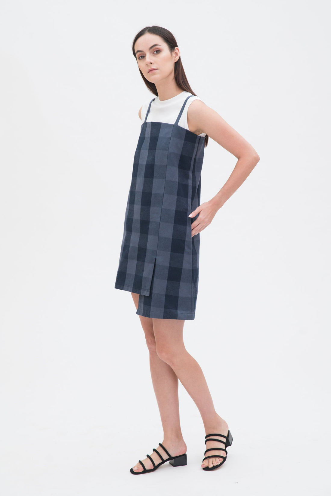 A woman with long dark hair is wearing a navy check dress with a white top underneath and  black sandals