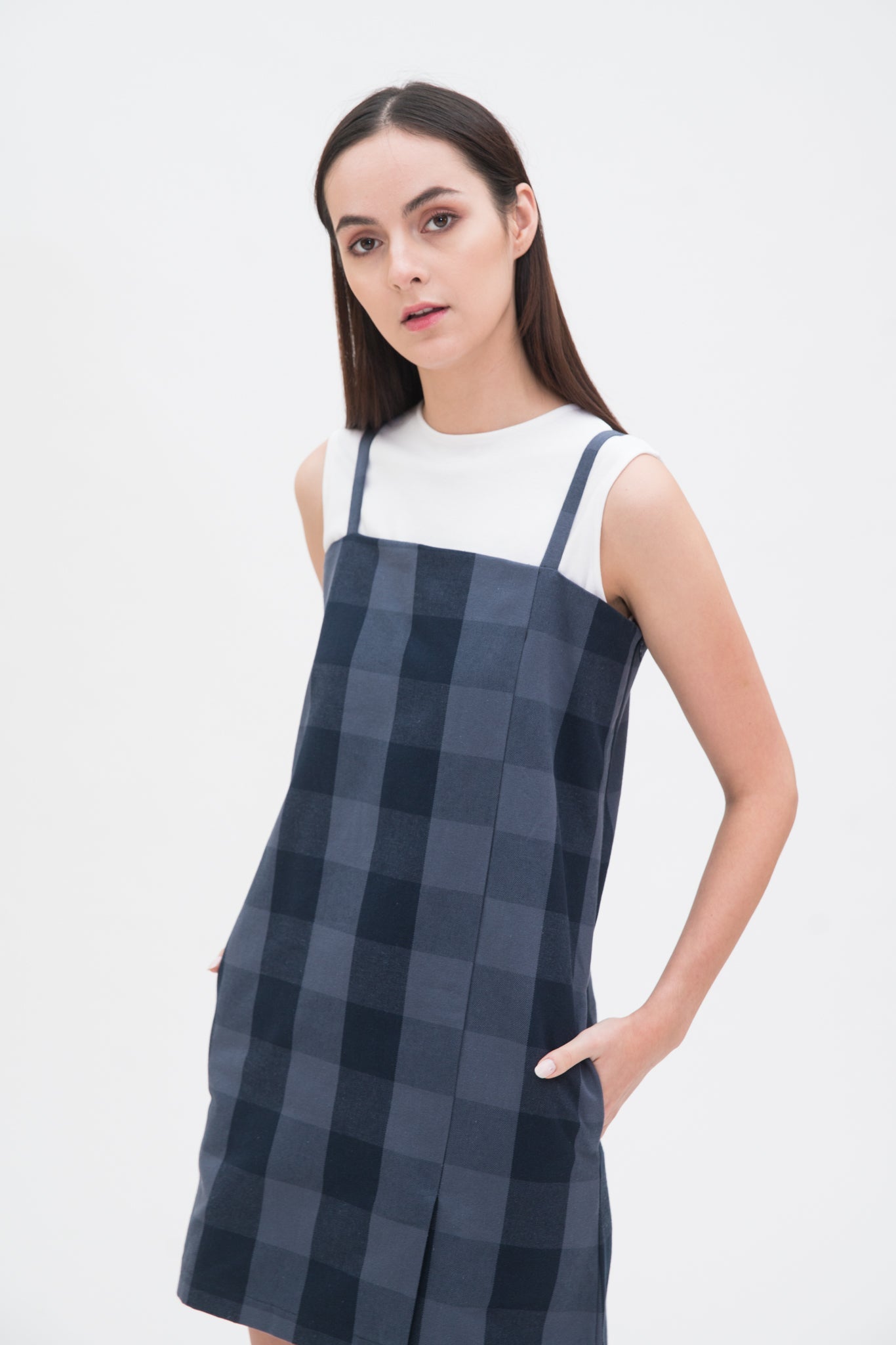 A woman with long dark hair is wearing a navy check dress with a white top underneath and  black sandals