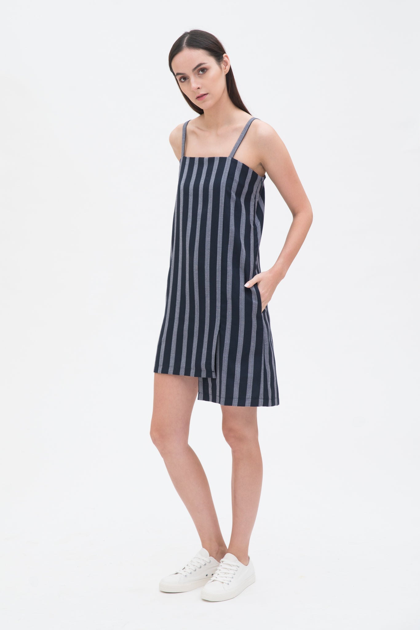 A woman with long brown hair is wearing a navy striped strappy dress. She has white trainers on.