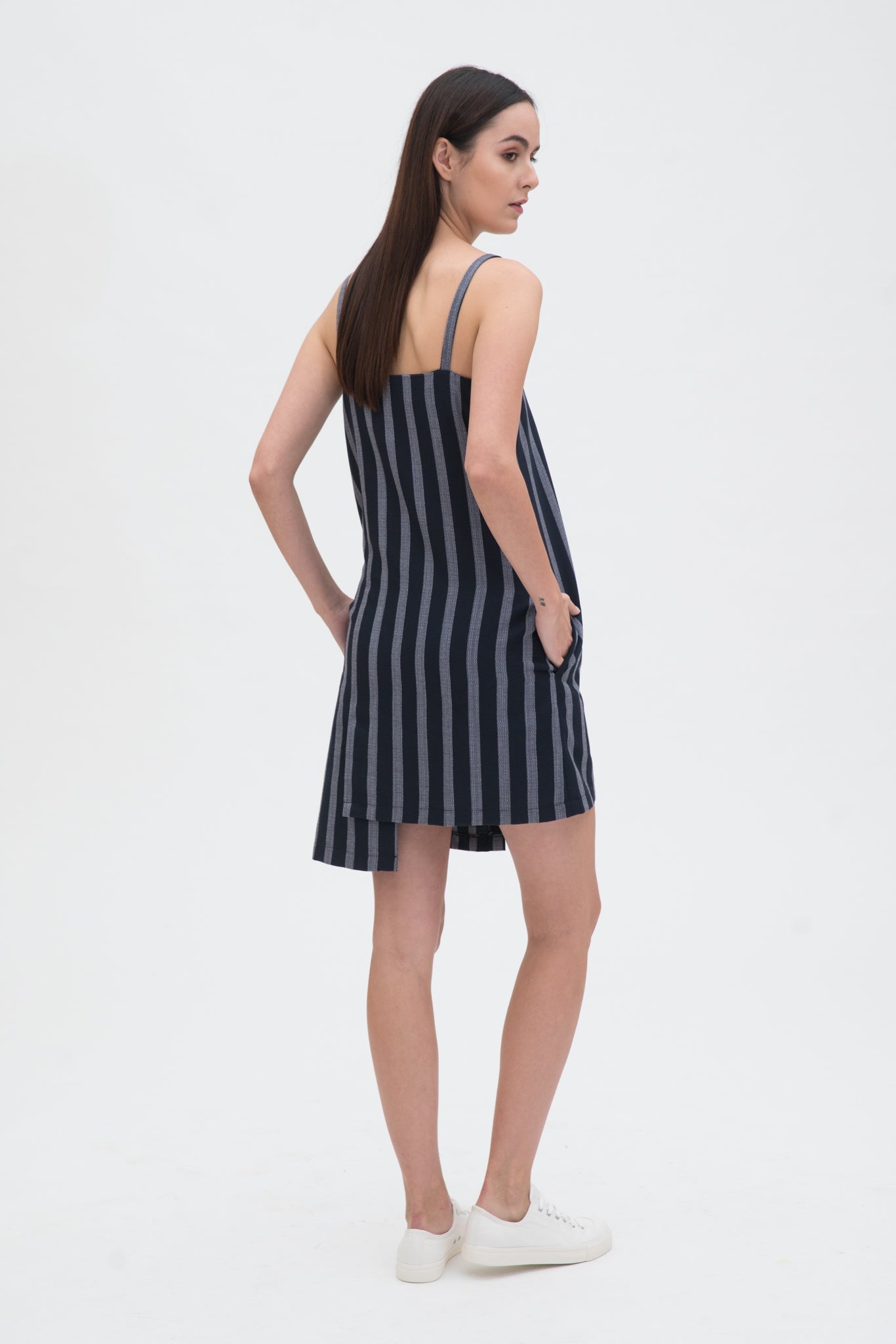 A woman with long brown hair is wearing a navy striped strappy dress. She has white trainers on.