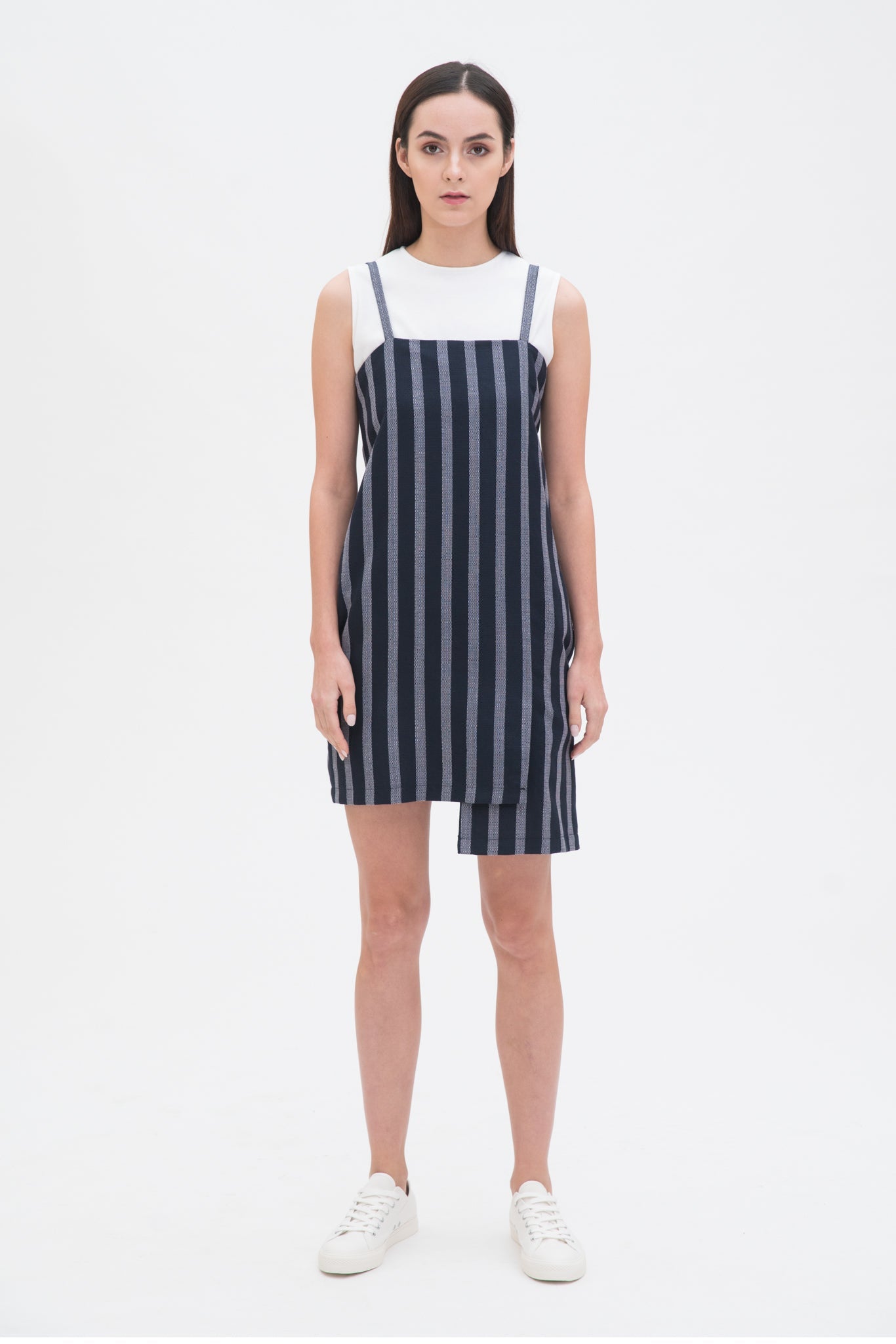 A woman with long brown hair is wearing a navy striped scrappy dress with a white top underneath. She has white trainers on.