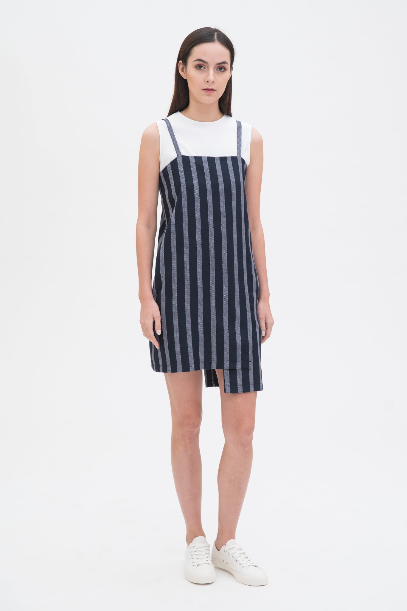 A woman with long brown hair is wearing a navy striped scrappy dress with a white top underneath. She has white trainers on.