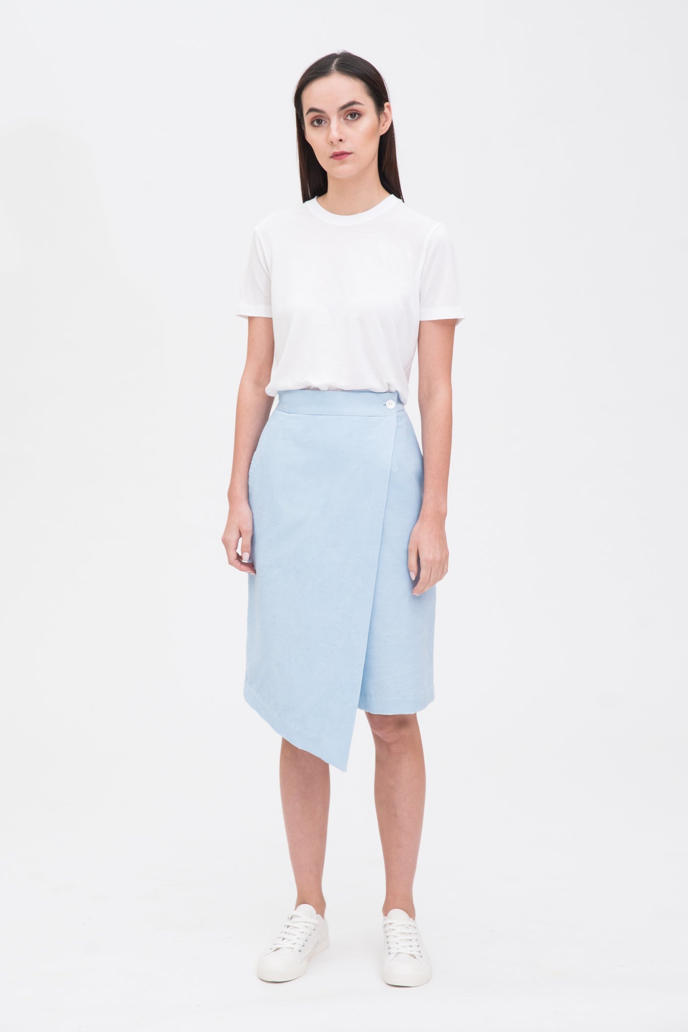 A woman with long dark hair is wearing a white t-shirt, light blue asymmetric skirt and white tennis shoes.