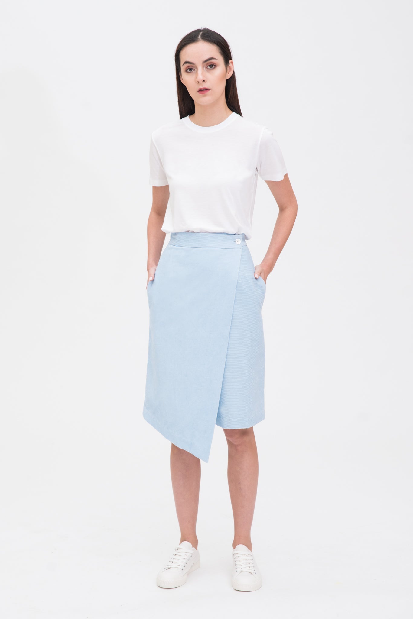 A woman with long dark hair is wearing a white t-shirt, light blue asymmetric skirt and white tennis shoes.