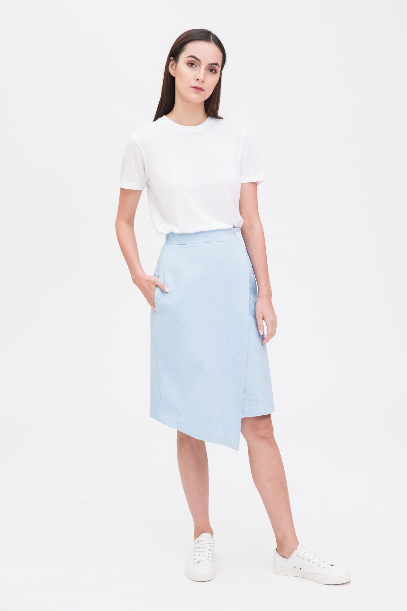 A woman with long dark hair is wearing a white t-shirt, light blue asymmetric skirt and white tennis shoes.