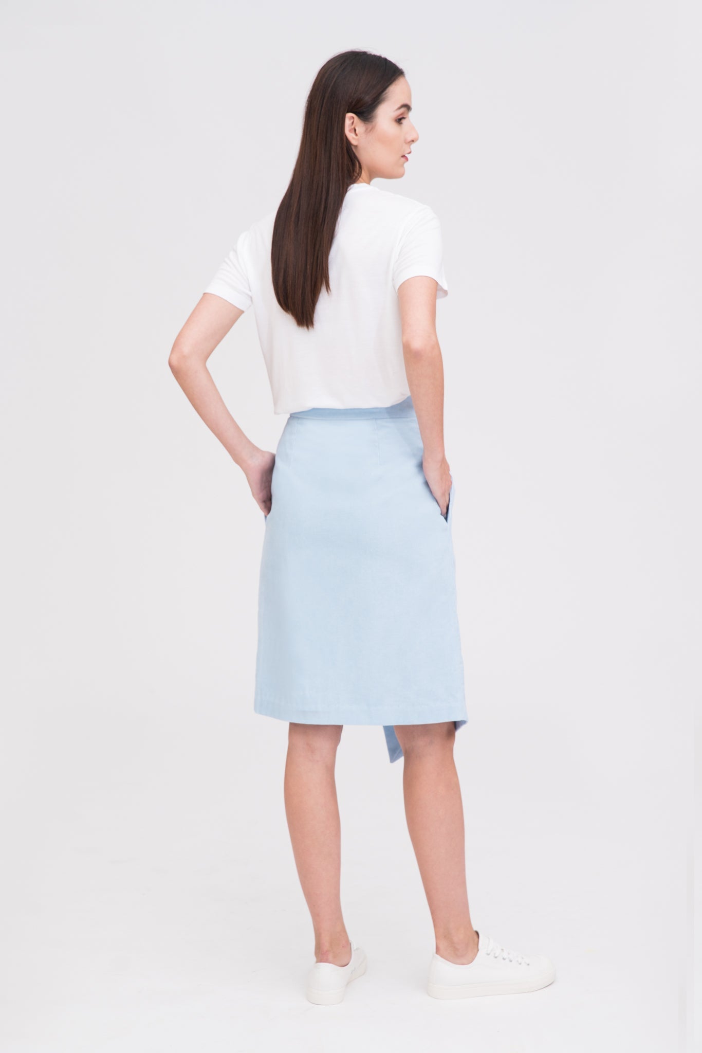 A woman with long dark hair is wearing a white t-shirt, light blue asymmetric skirt and white tennis shoes.