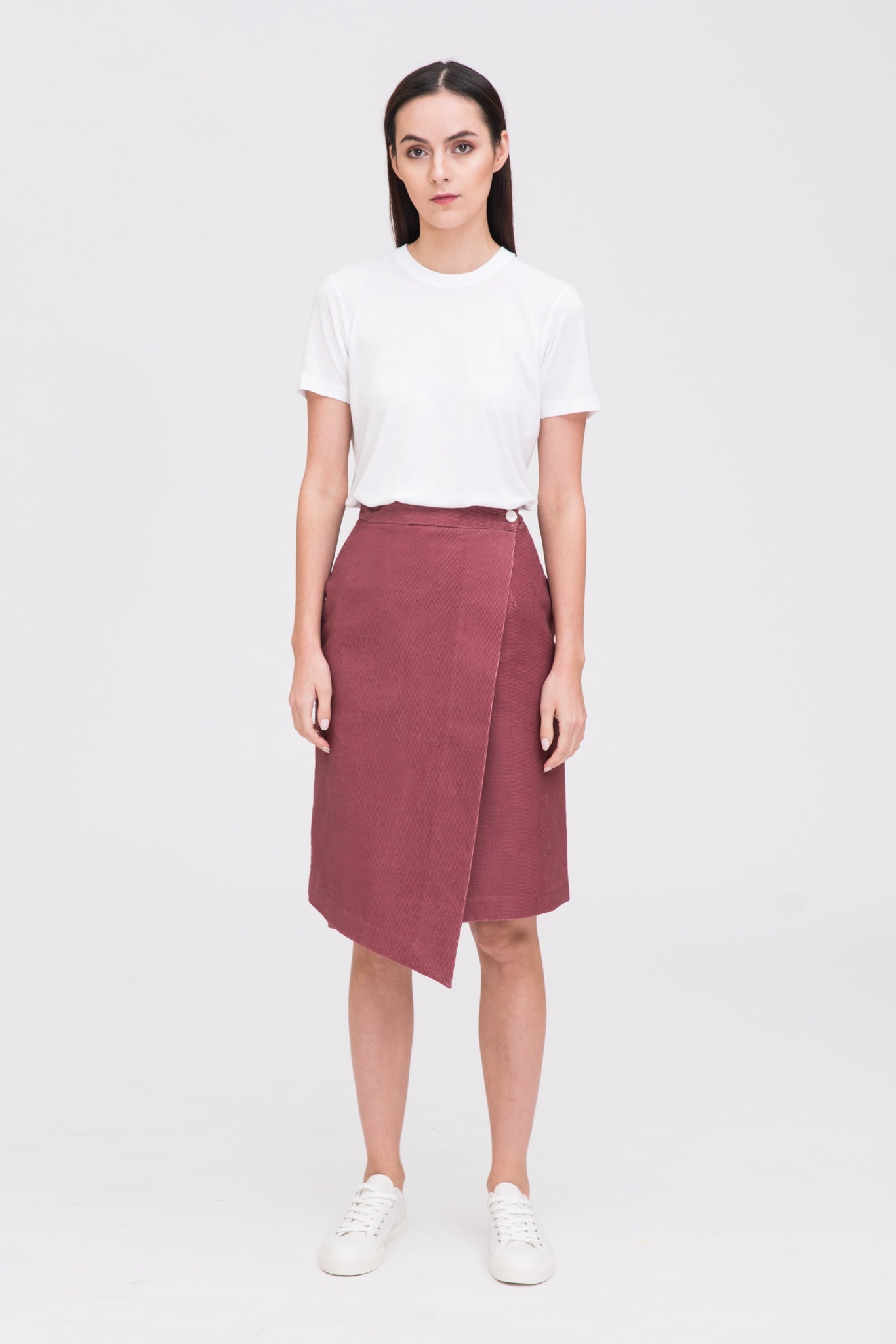 A woman with long dark hair is wearing a white t-shirt and dusty pink asymmetric skirt. She is wearing white tennis shoes.