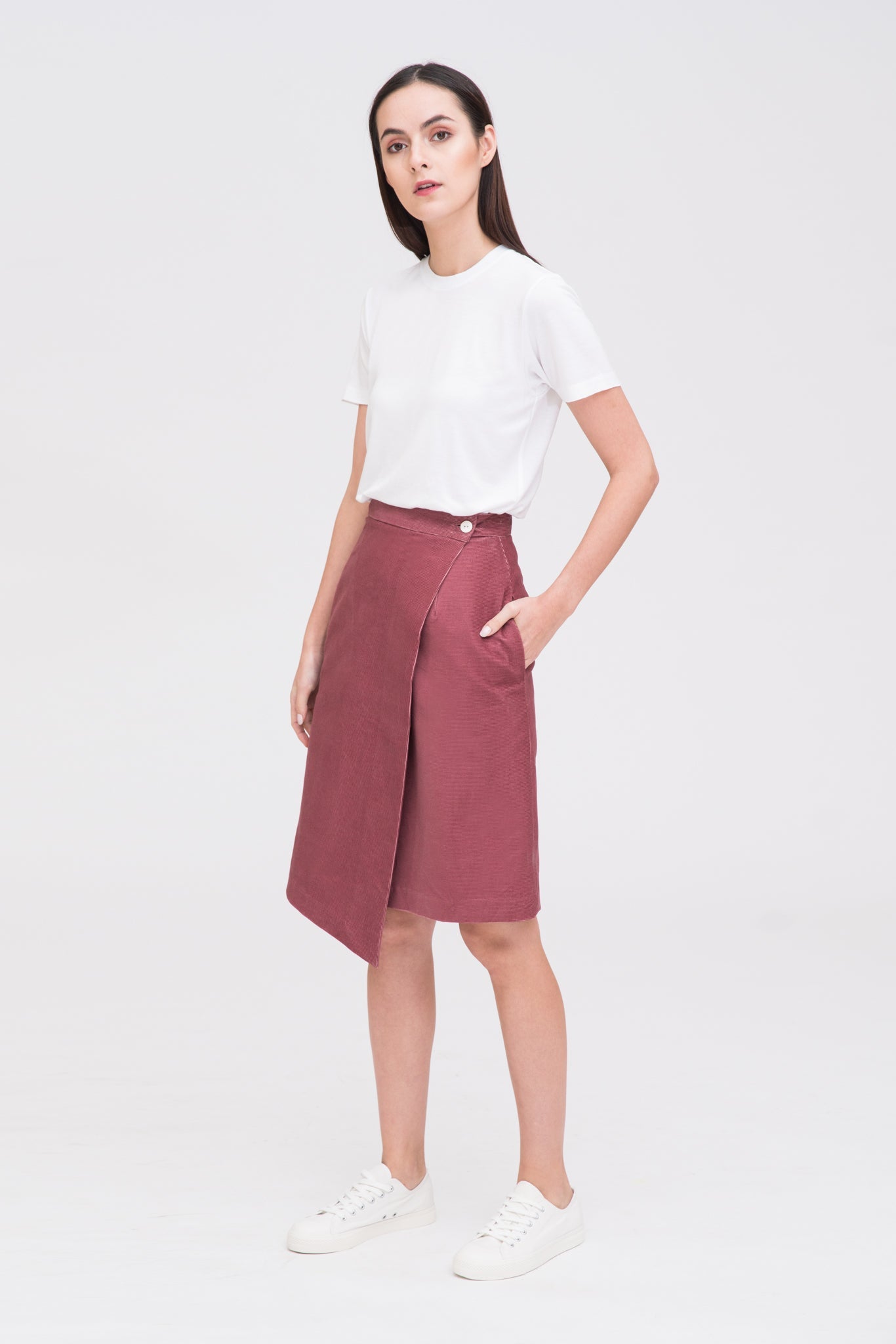 A woman with long dark hair is wearing a white t-shirt and dusty pink asymmetric skirt. She is wearing white tennis shoes.