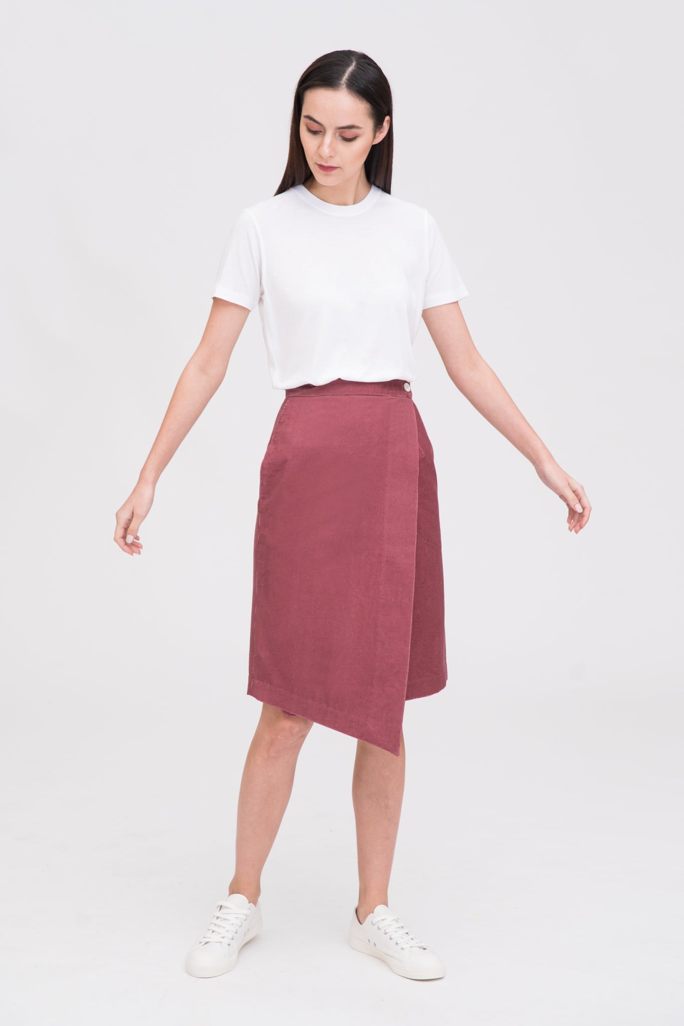A woman with long dark hair is wearing a white t-shirt and dusty pink asymmetric skirt. She is wearing white tennis shoes.