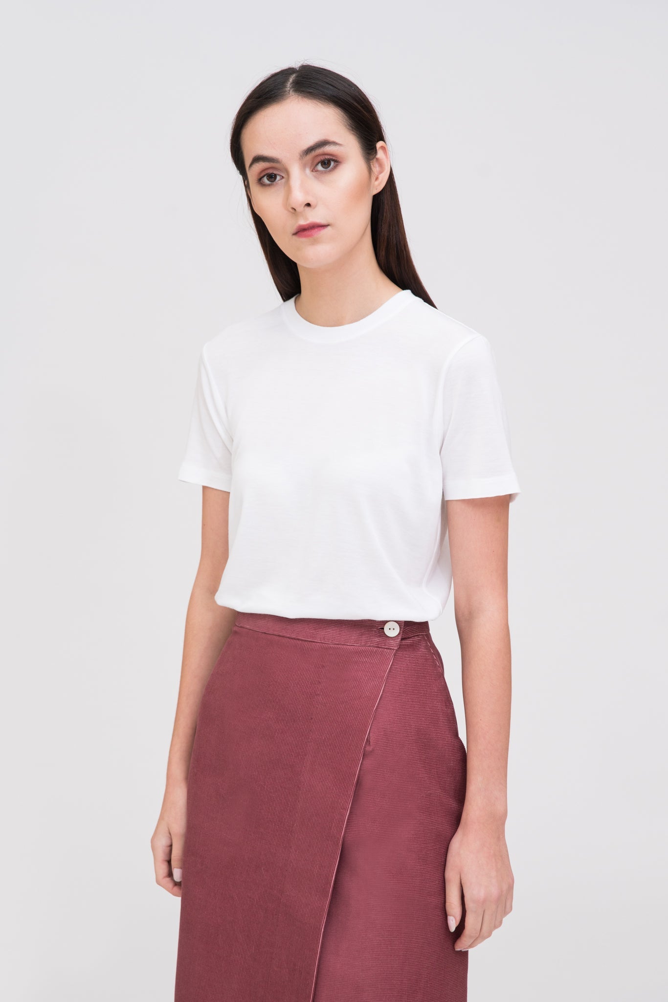 A woman with long dark hair is wearing a white t-shirt and dusty pink asymmetric skirt. She is wearing white tennis shoes.
