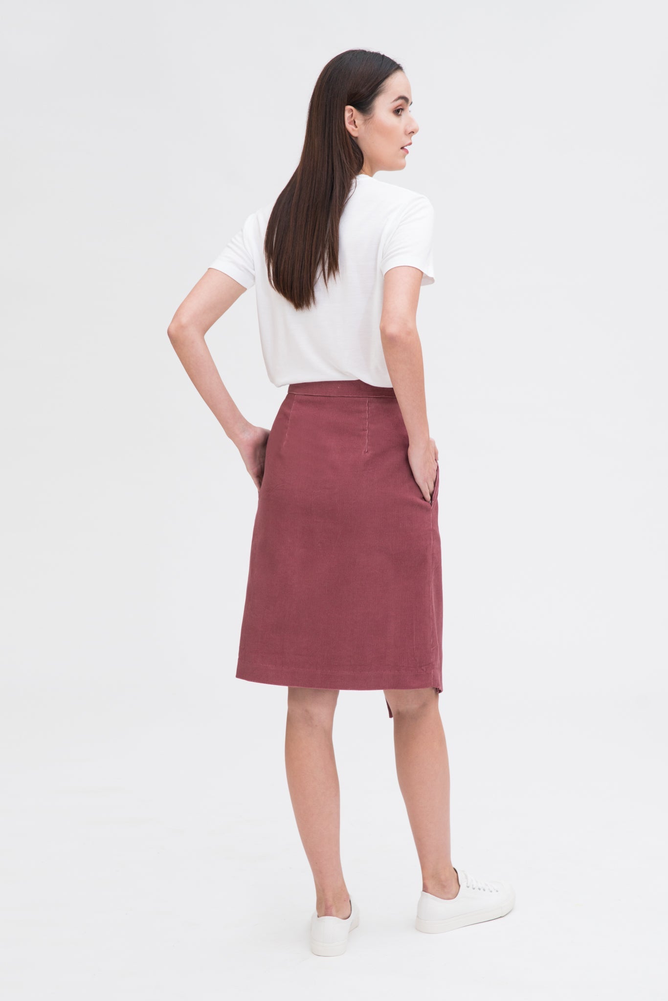 A woman with long dark hair is wearing a white t-shirt, dusty pink asymmetric skirt and white tennis shoes.