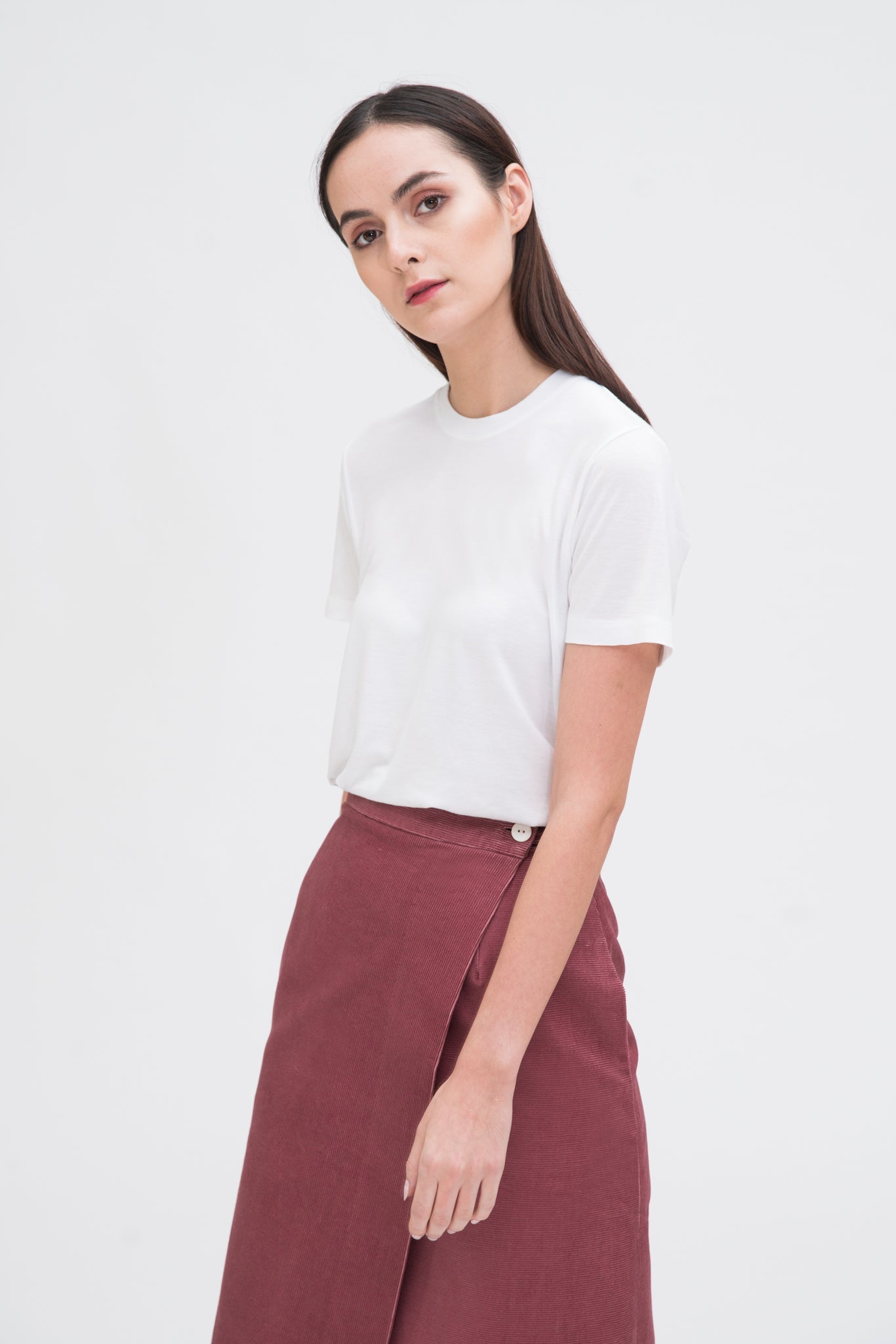 A woman with long dark hair is wearing a white t-shirt and dusty pink asymmetric skirt.
