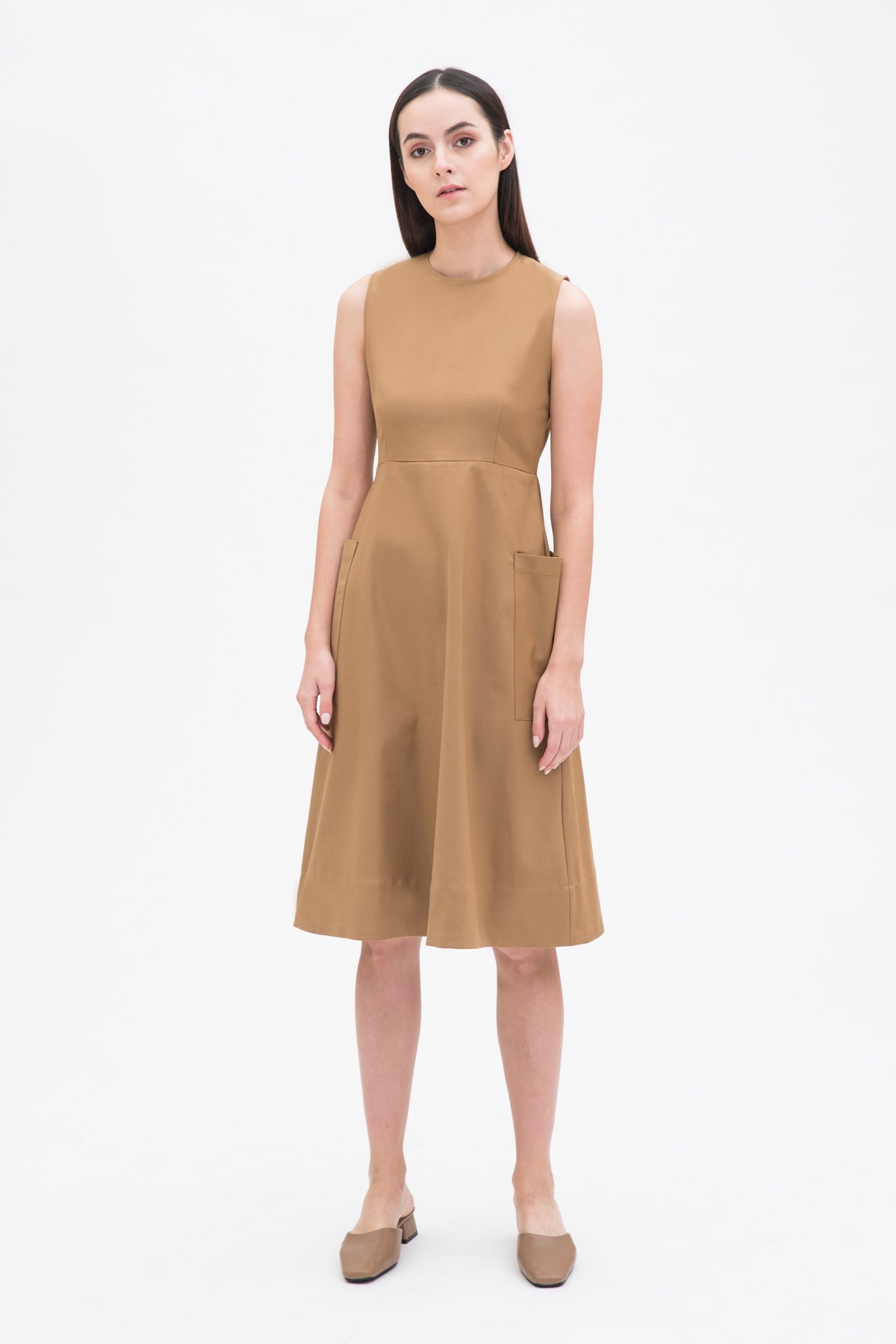 A woman with long dark hair is wearing a knee length camel dress with pockets. She is wearing camel leather shoes.