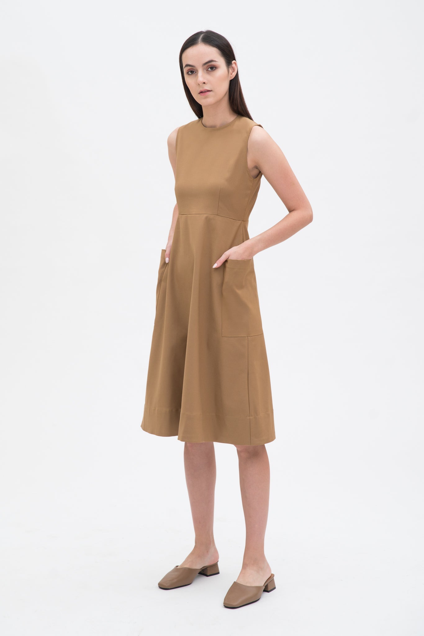 A woman with long dark hair is wearing a knee length camel dress with pockets. She is wearing camel leather shoes.