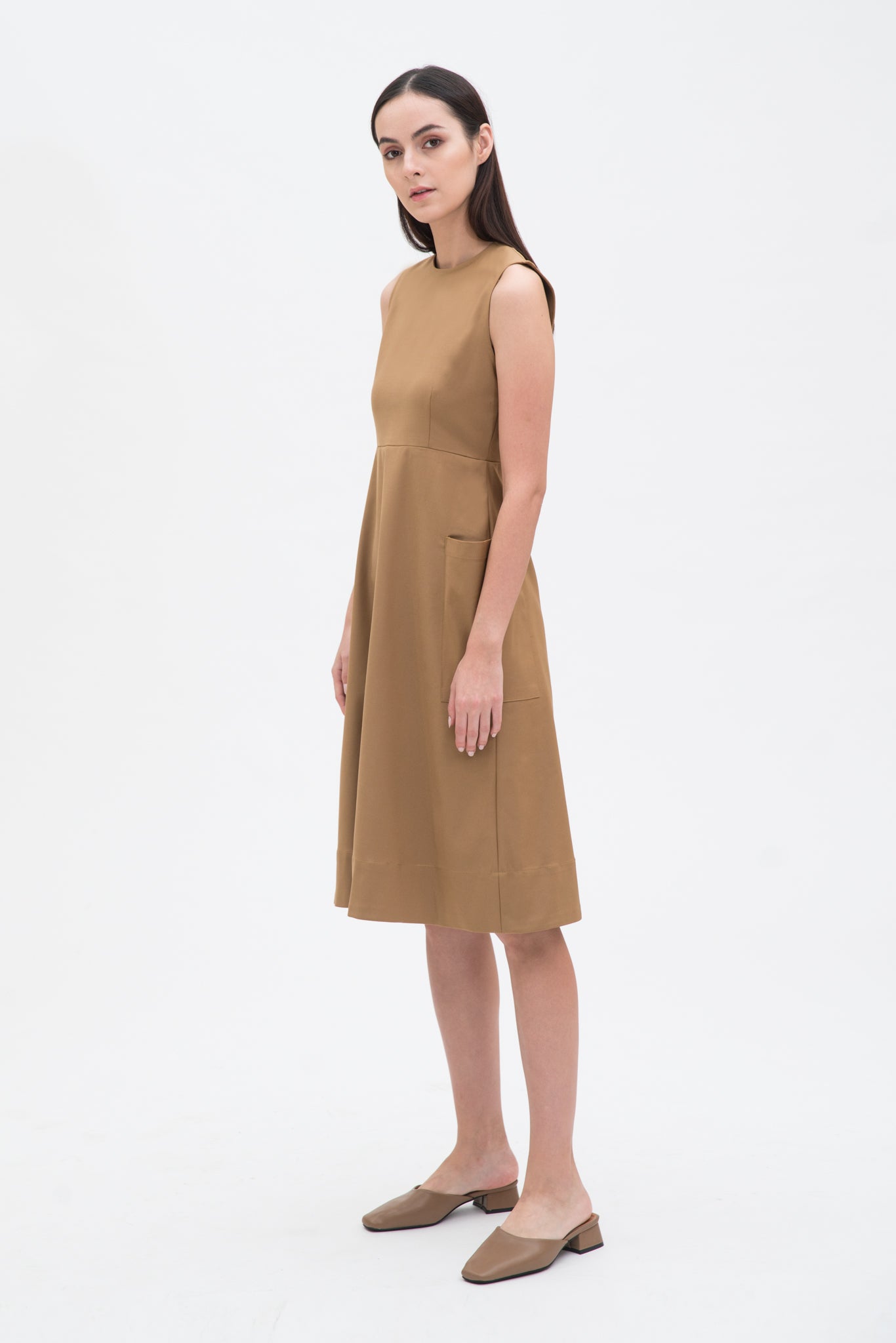 A woman with long dark hair is wearing a knee length camel dress with pockets. She is wearing camel leather shoes.