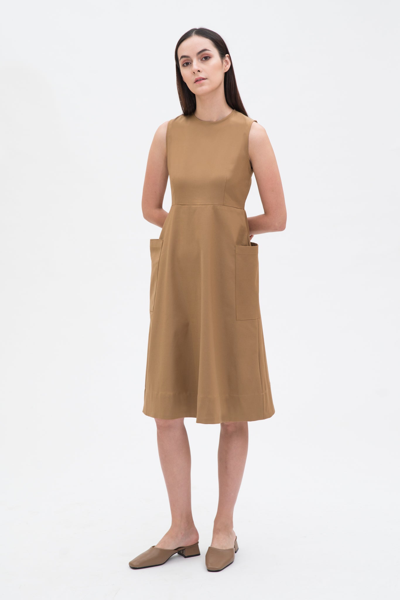 A woman with long dark hair is wearing a knee length camel dress with pockets. She is wearing camel leather shoes.