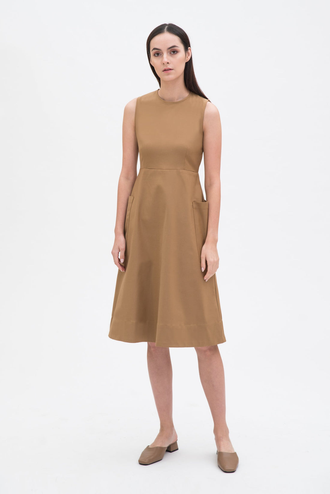 A woman with long dark hair is wearing a knee length camel dress with pockets. She is wearing camel leather shoes.
