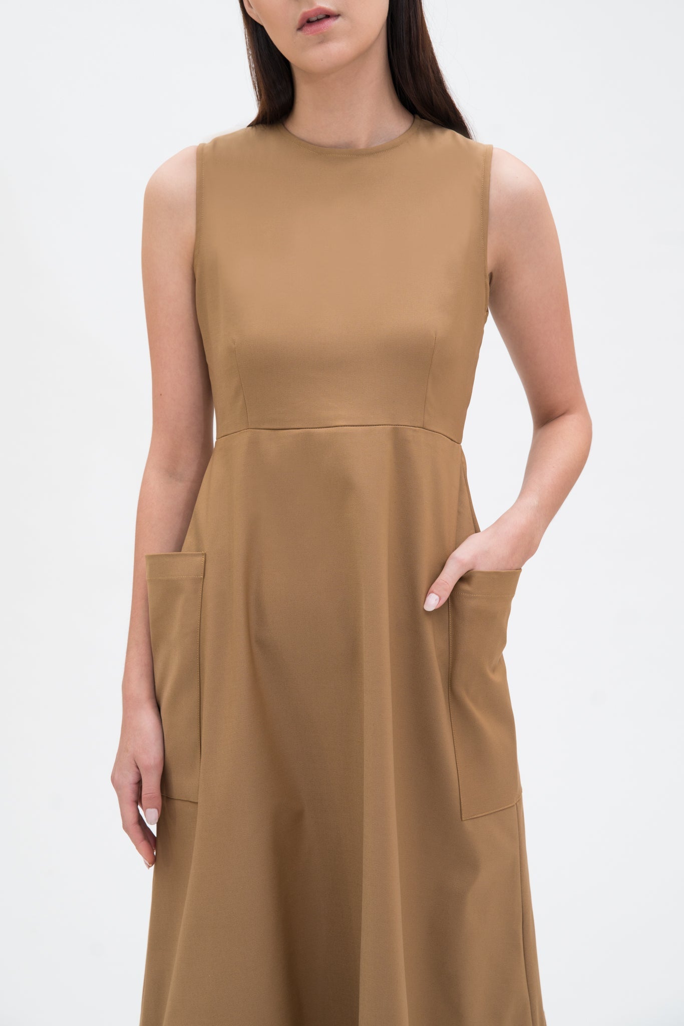 A woman with long dark hair is wearing a knee length camel dress with pockets.