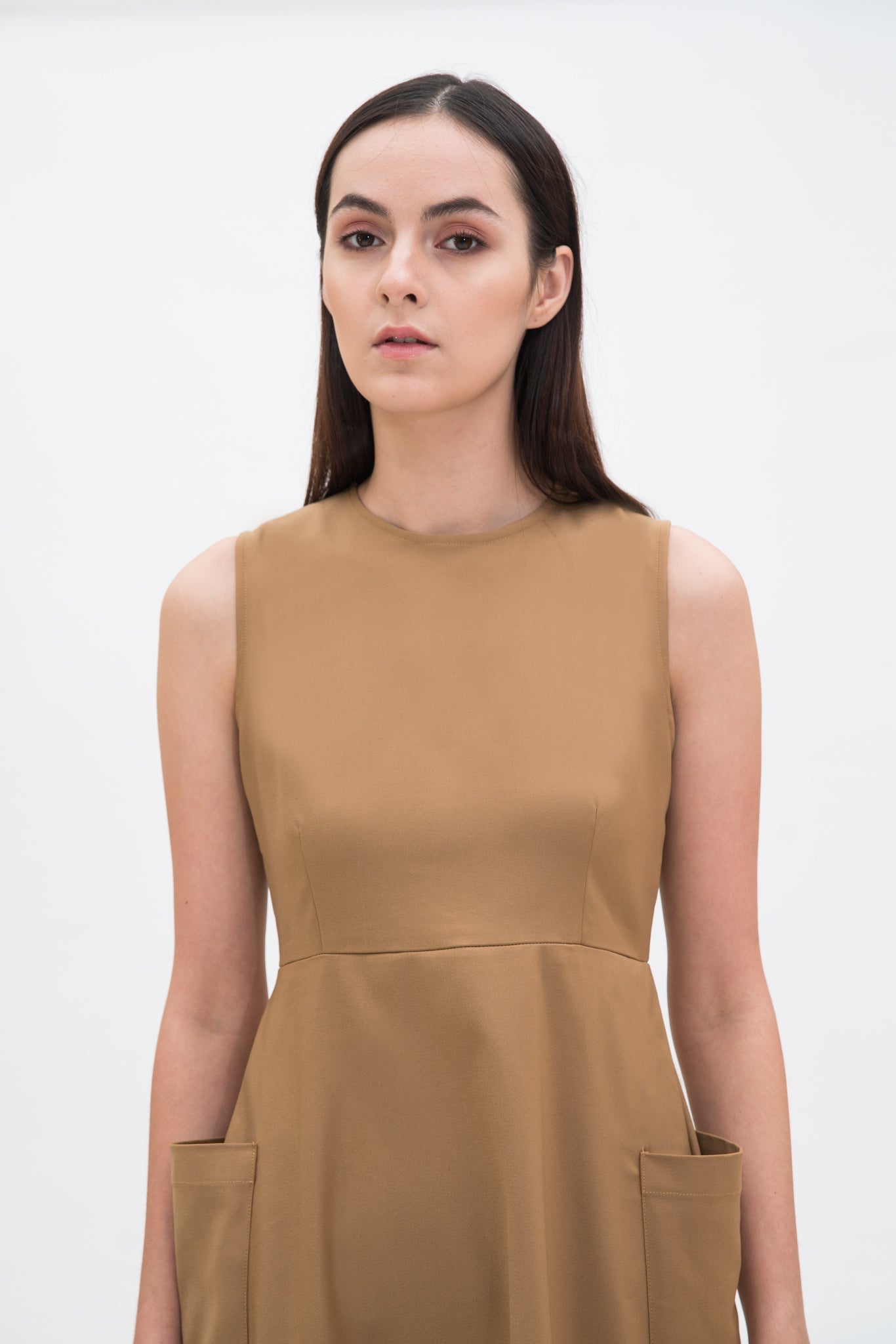 A woman with long dark hair is wearing a knee length camel dress with pockets.