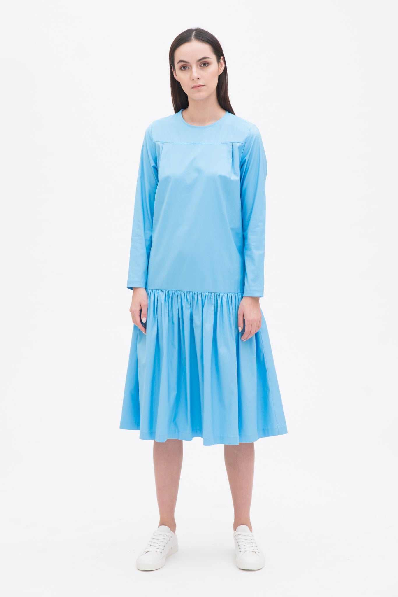 A woman with long brown hair is wearing a drop waist blue dress with ruffle hem. She is also wearing white tennis shoes.