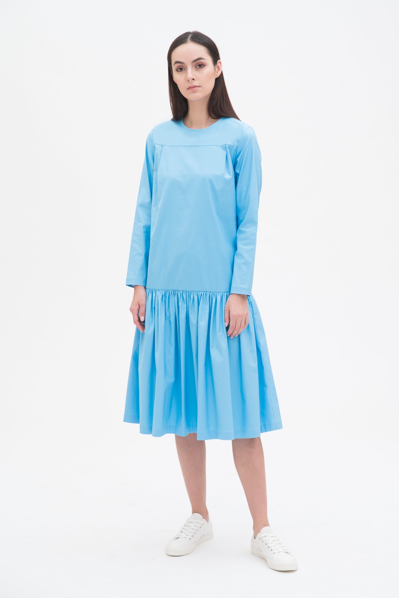 A woman with long brown hair is wearing a drop waist blue dress with ruffle hem. She is also wearing white tennis shoes.