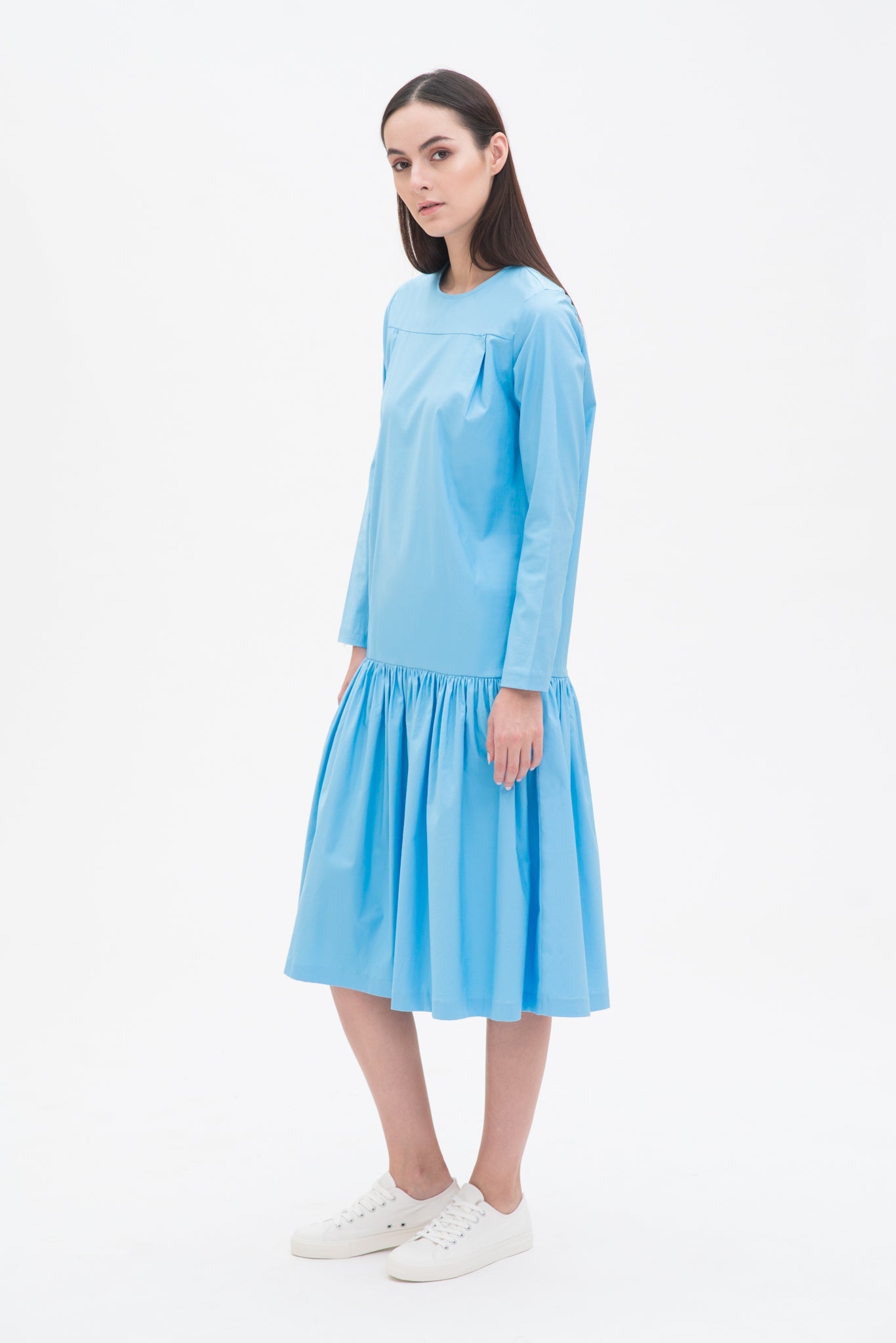 A woman with long brown hair is wearing a drop waist blue dress with ruffle hem. She is also wearing white tennis shoes.
