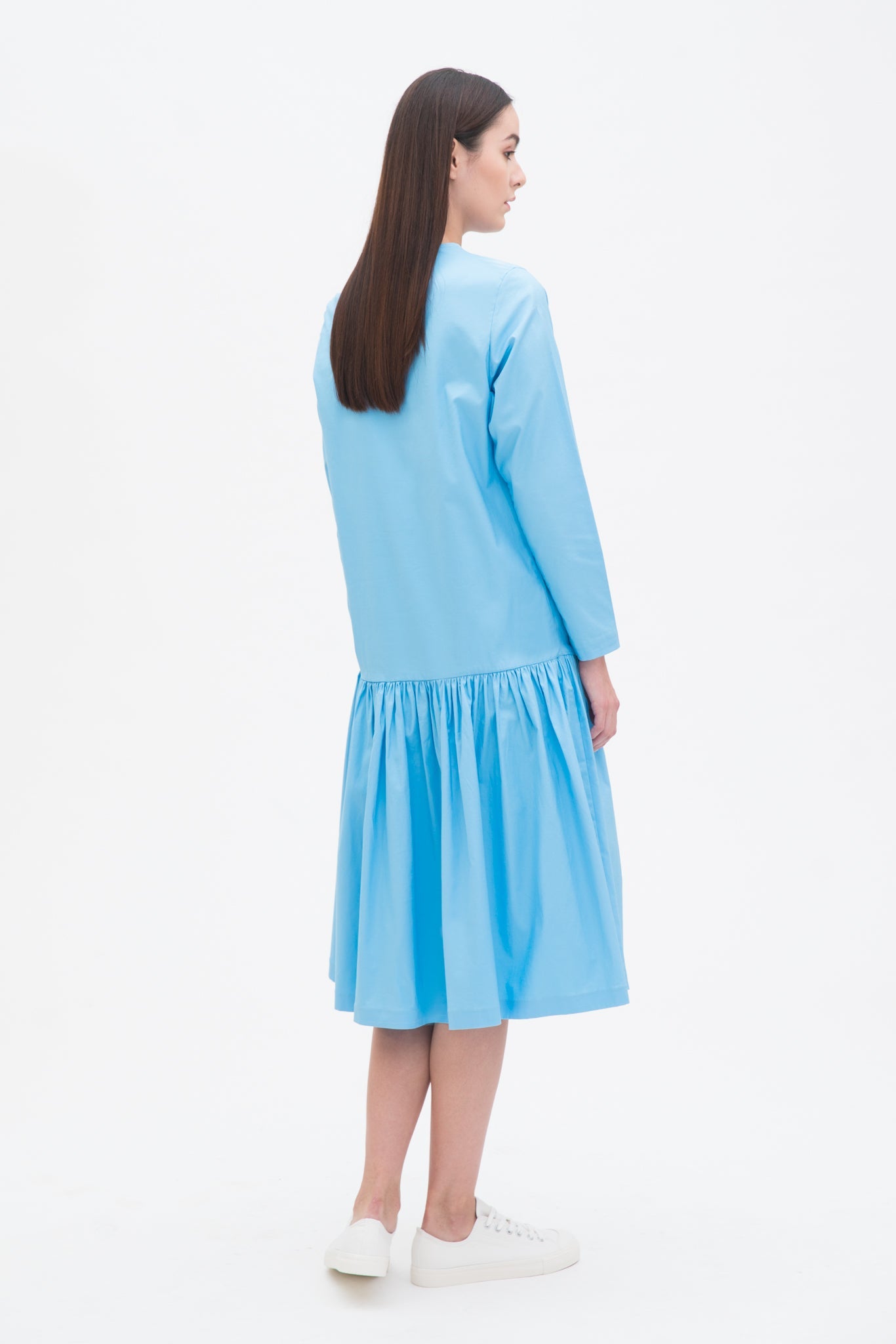 A woman with long brown hair is wearing a drop waist blue dress with ruffle hem. She is also wearing white tennis shoes.