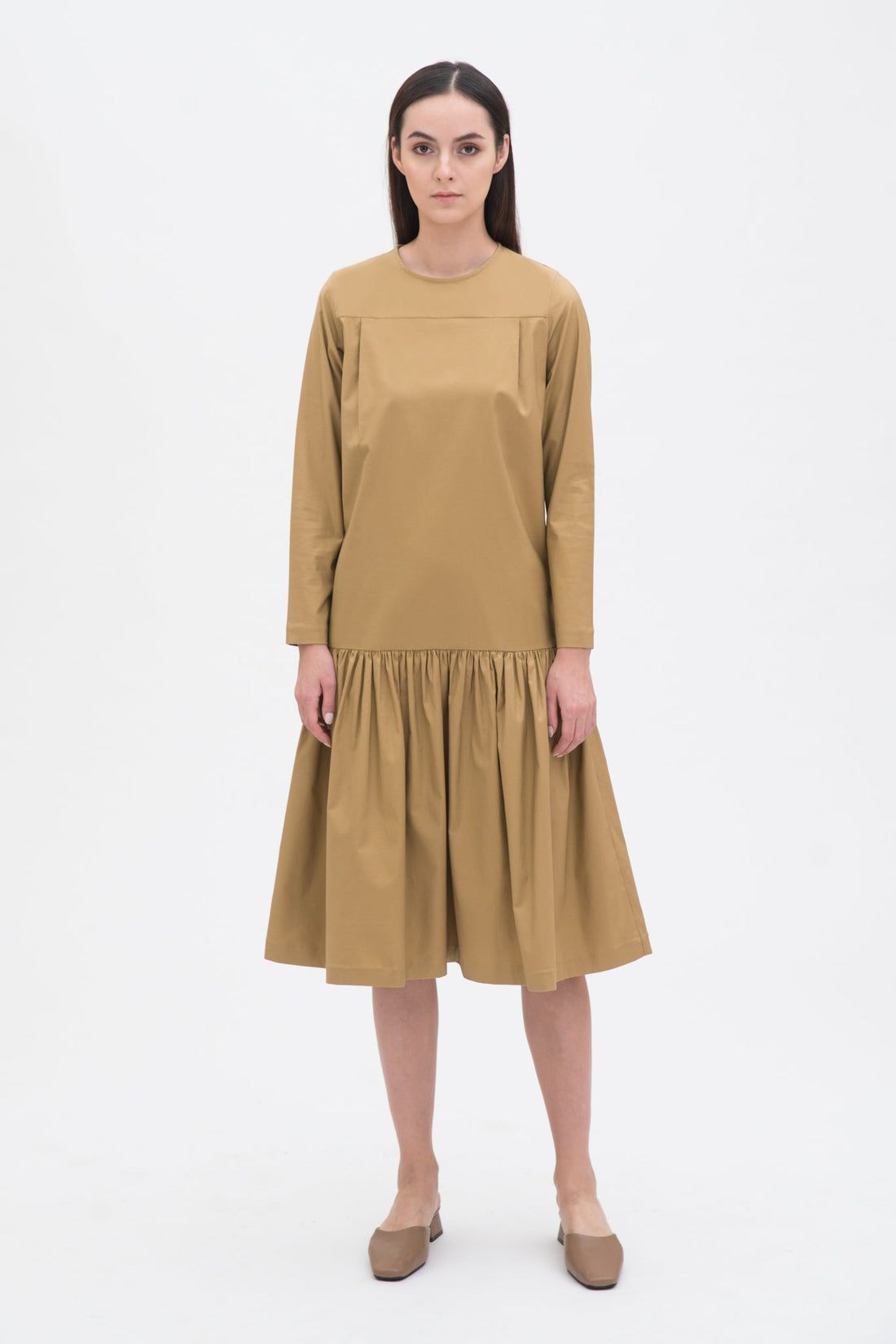 A woman with long brown hair is wearing a drop waist camel dress with ruffle hem. She is also wearing beige leather shoes.