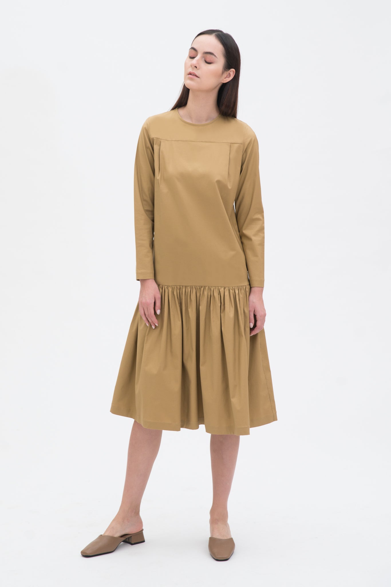 A woman with long brown hair is wearing a drop waist camel dress with ruffle hem. She is also wearing beige leather shoes.