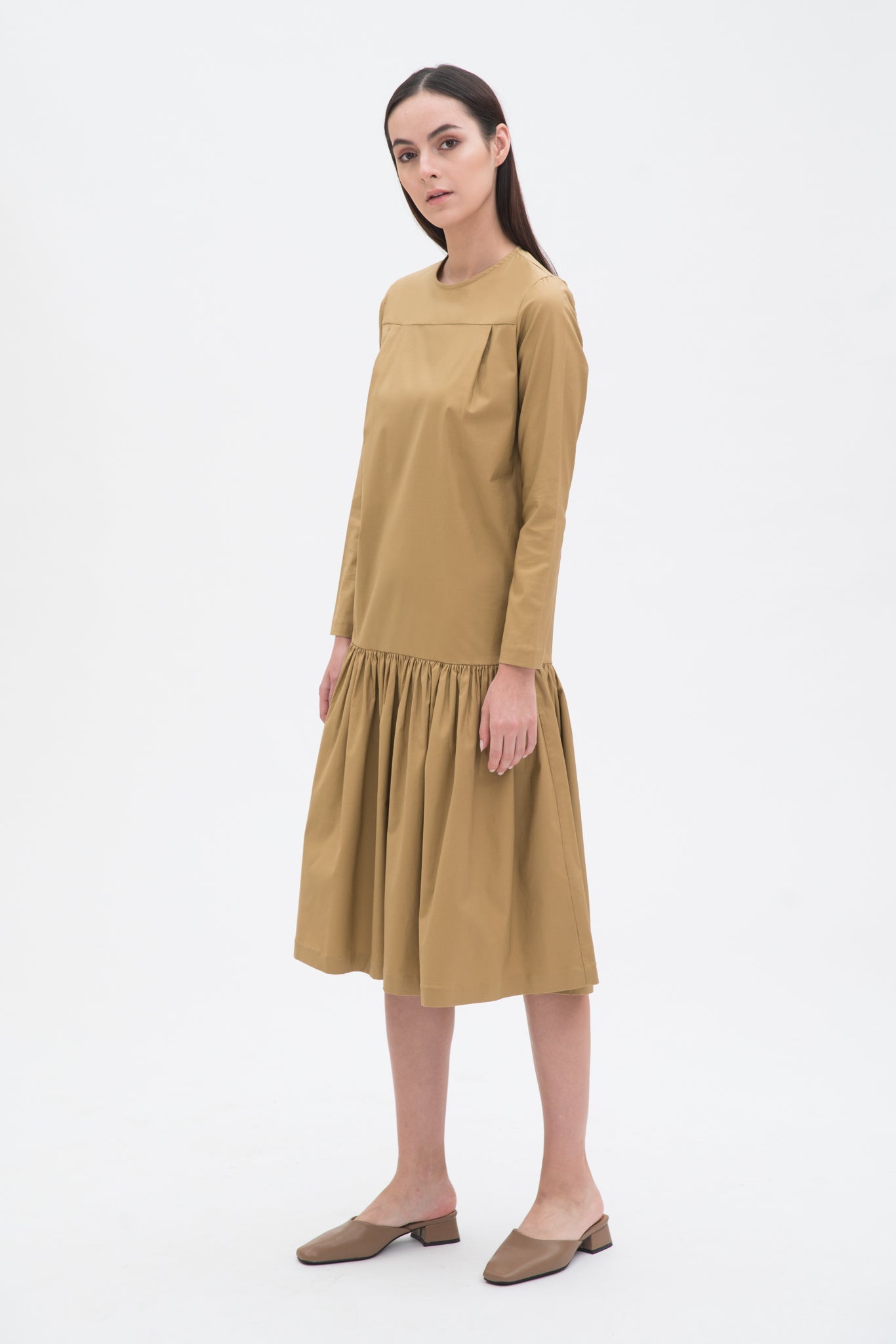 A woman with long brown hair is wearing a drop waist camel dress with ruffle hem. She is also wearing beige leather shoes.