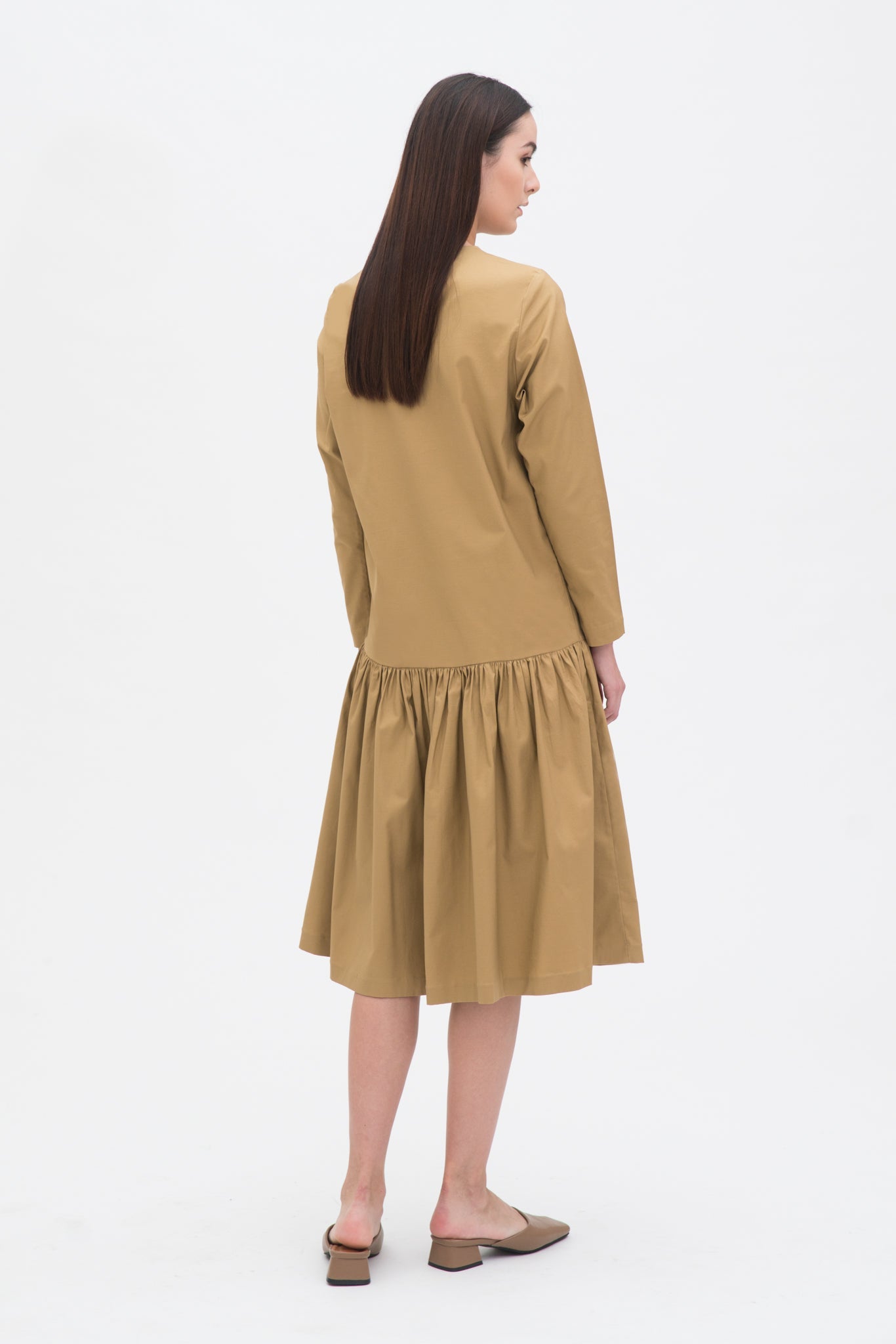 A woman with long brown hair is wearing a drop waist camel dress with ruffle hem. She is also wearing beige leather shoes.