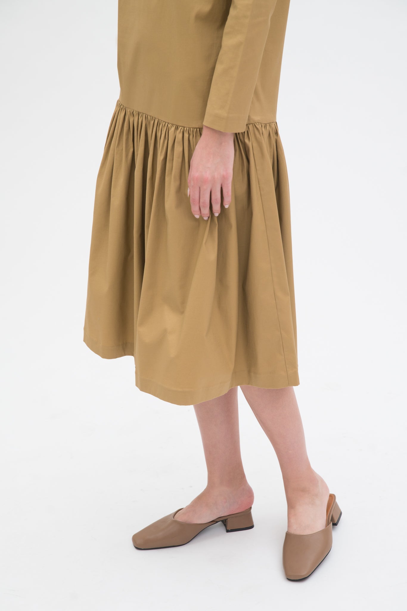 A woman with long brown hair is wearing a drop waist camel dress with ruffle hem. She is also wearing beige leather shoes.