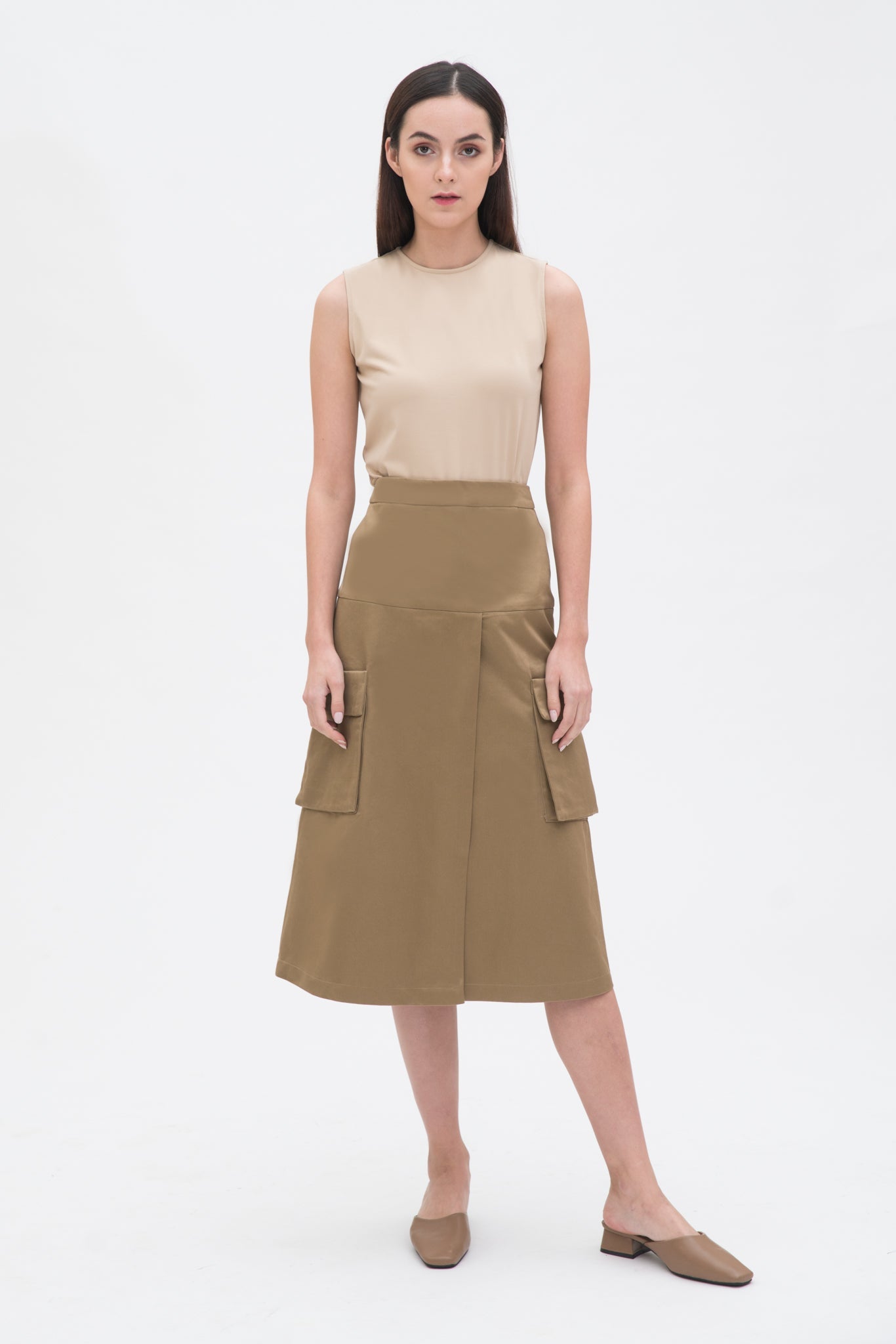 A woman with long dark hair is wearing a beige tank top and a knee length camel skirt with large pockets. She is also wearing camel leather shoes.