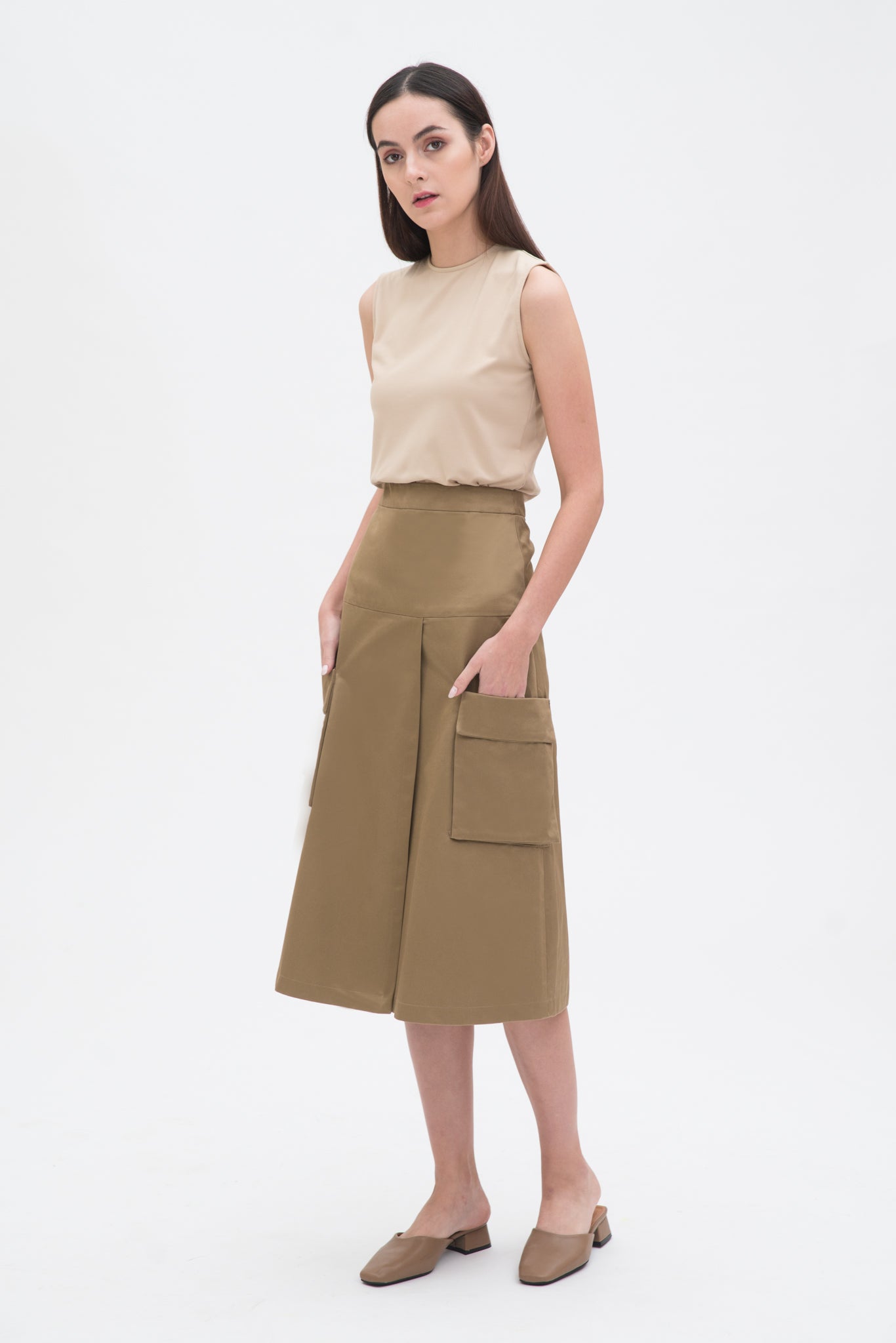 A woman with long dark hair is wearing a beige tank top and a knee length camel skirt with large pockets. She is also wearing camel leather shoes.