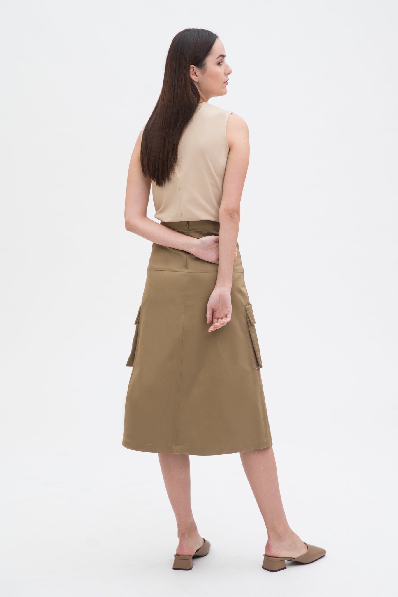 A woman with long dark hair is wearing a beige tank top and a knee length camel skirt with large pockets. She is also wearing camel leather shoes.