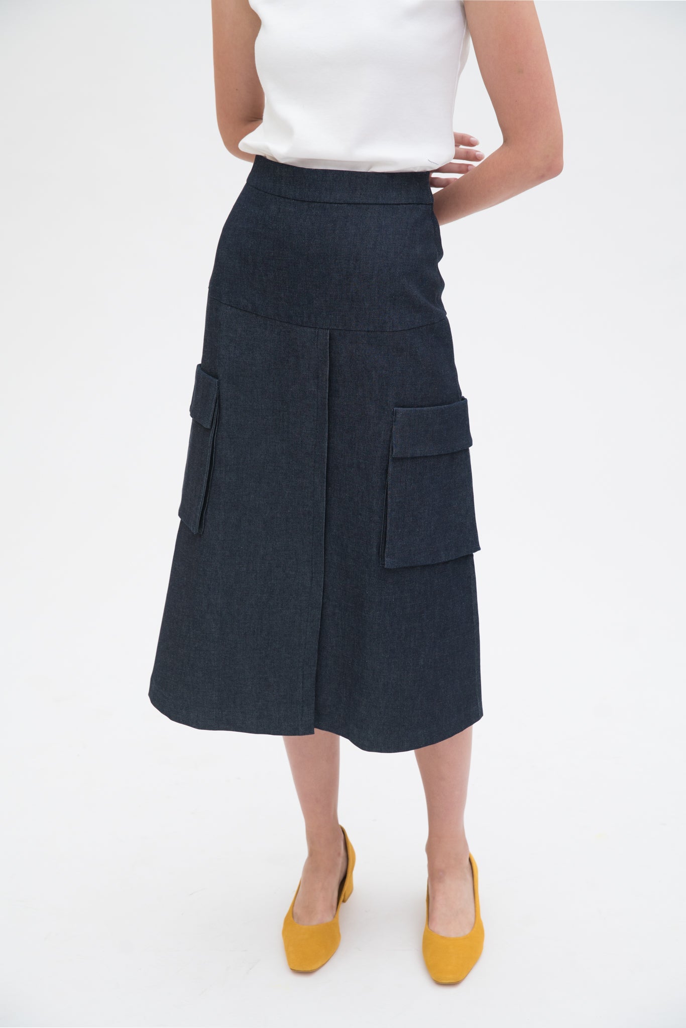 A woman with long dark hair is wearing a white tank top and a knee length navy skirt with large pockets. She is also wearing yellow leather shoes.