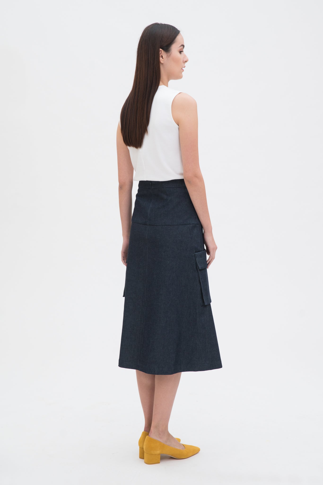 A woman with long dark hair is wearing a white tank top and a knee length navy skirt with large pockets. She is also wearing yellow leather shoes.