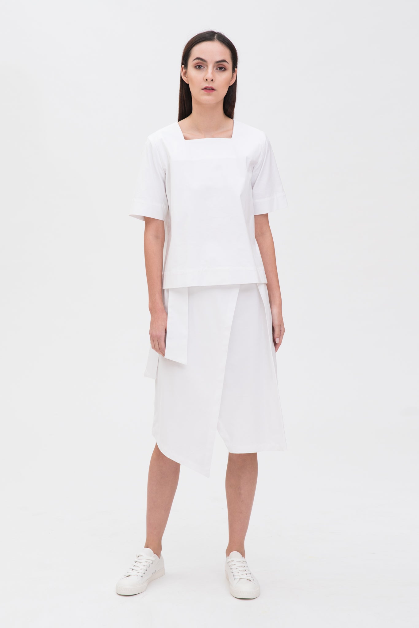 A woman with long dark hair is wearing a navy t-shirt and asymmetric white skirt  with white shoes