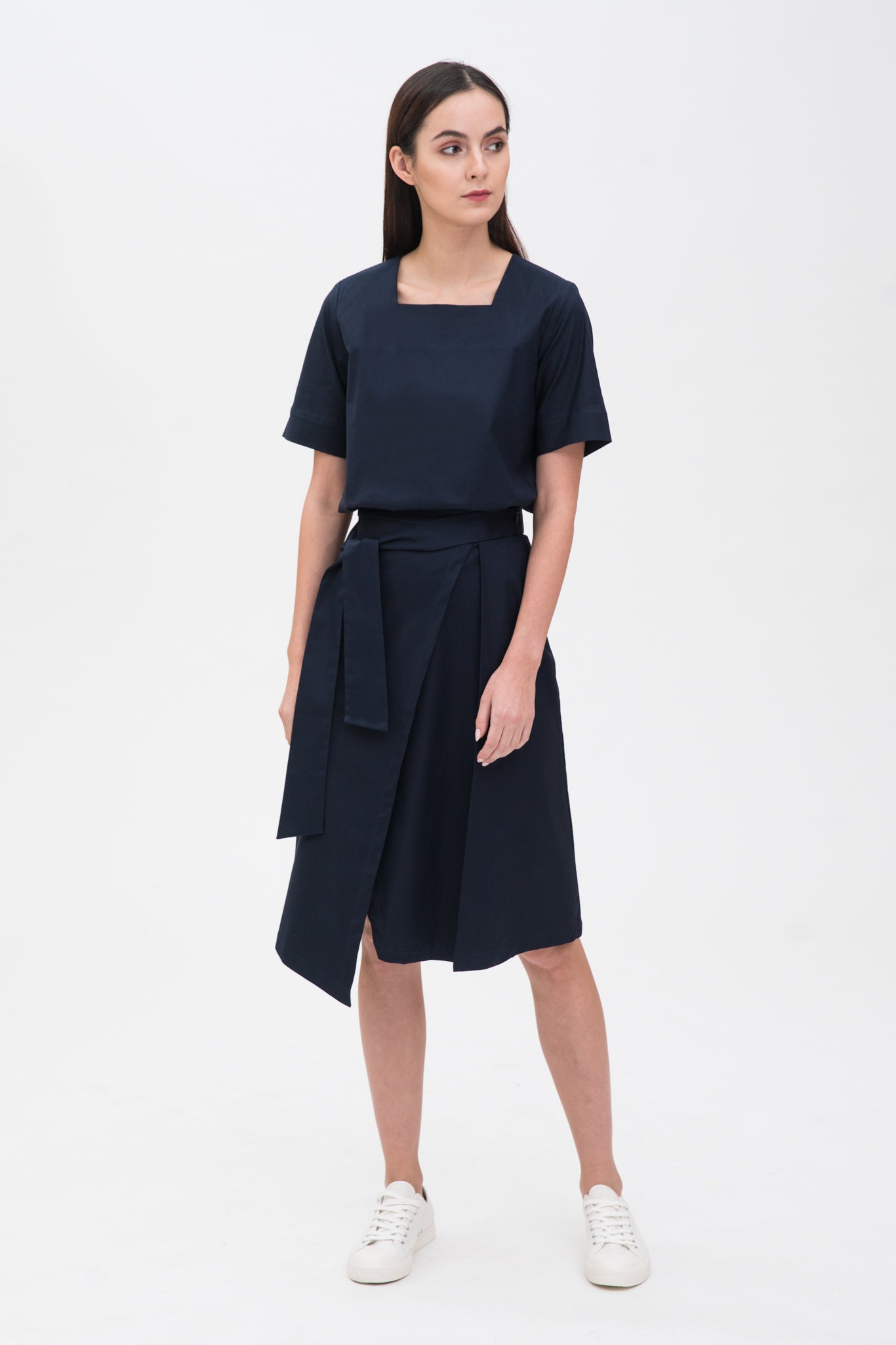 A woman with long dark hair is wearing a navy t-shirt and asymmetric navy skirt  with white shoes