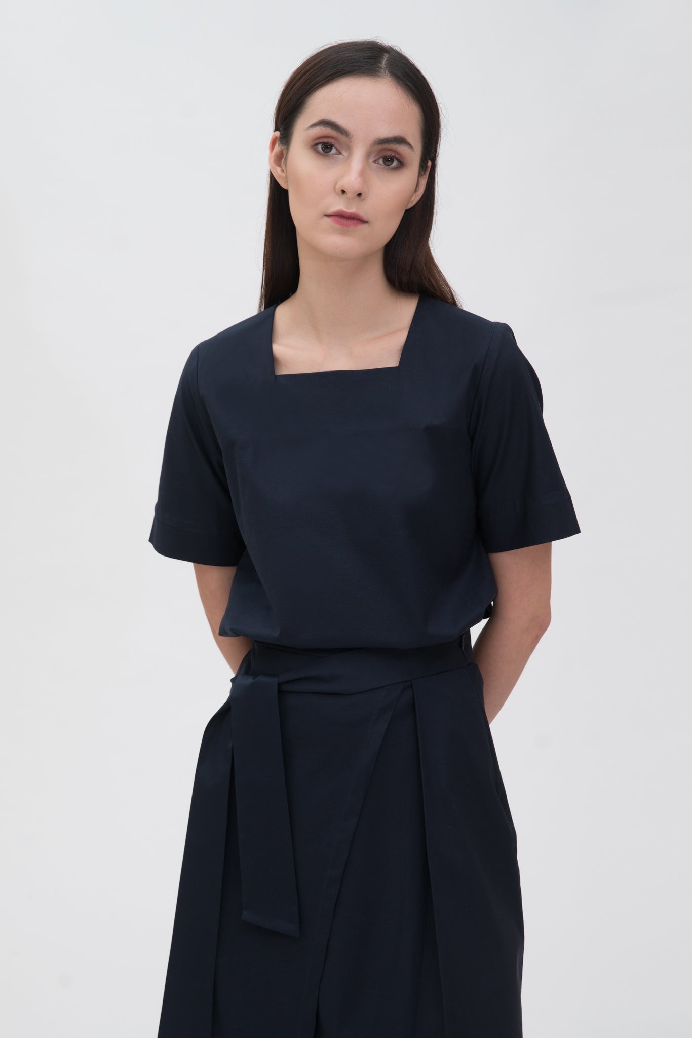 A woman with long dark hair is wearing a navy t-shirt and asymmetric navy skirt