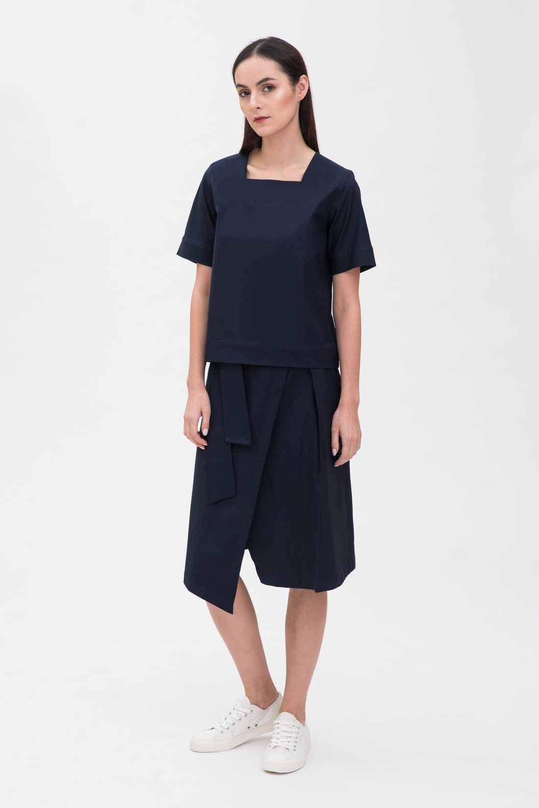 A woman with long dark hair is wearing a navy t-shirt and asymmetric navy skirt  with white shoes