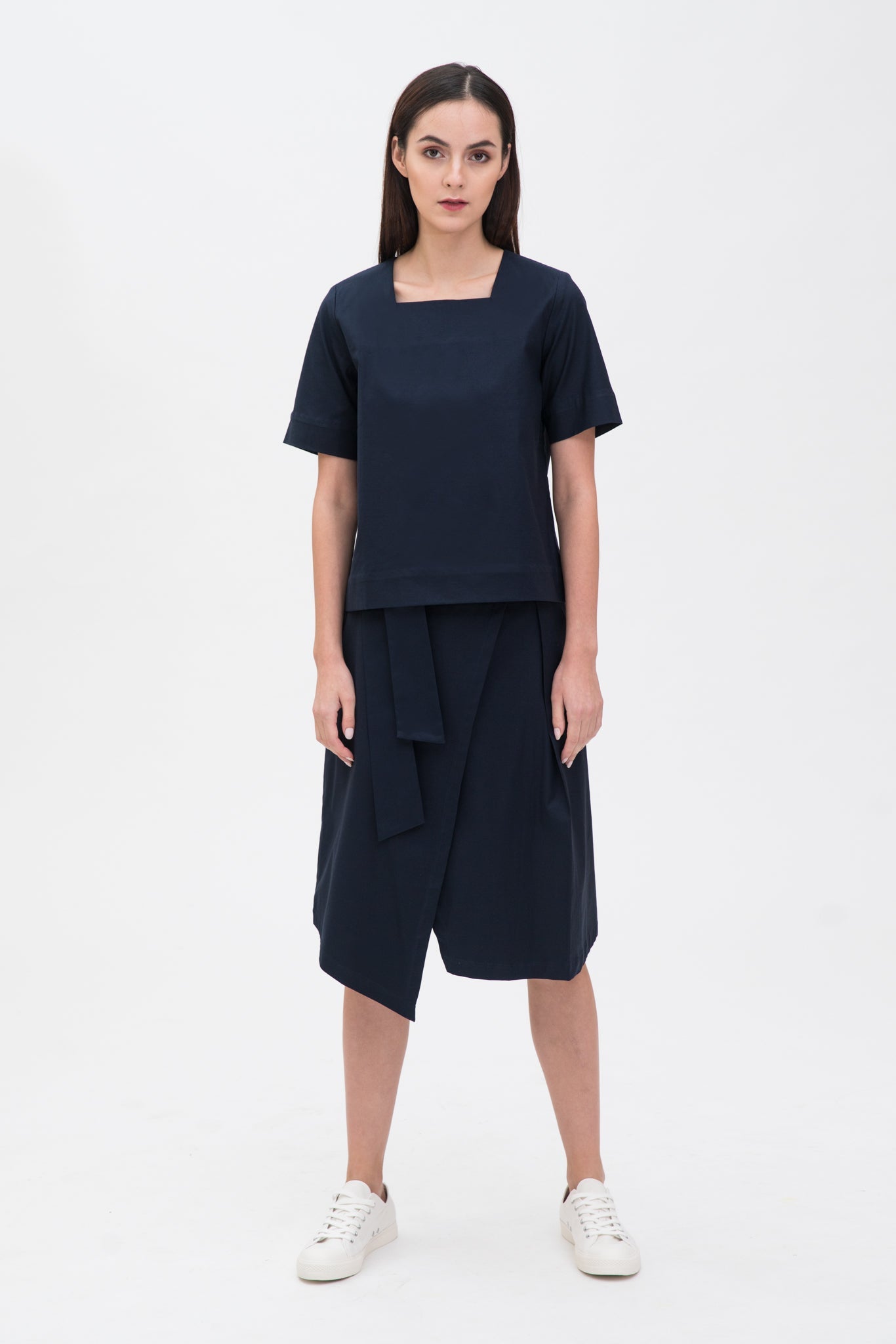 A woman with long dark hair is wearing a navy t-shirt and asymmetric navy skirt  with white shoes